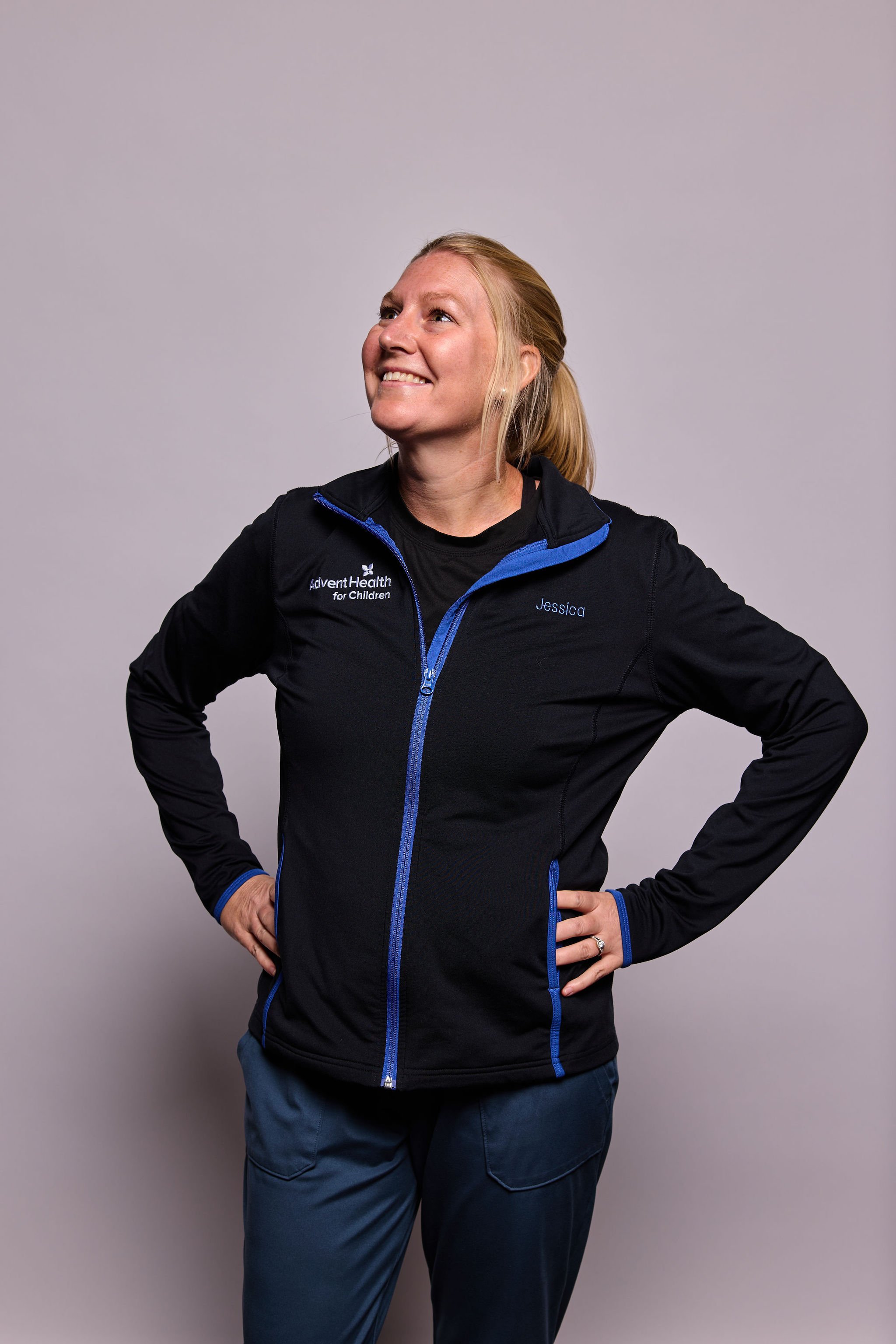 A woman with blonde hair tied back, smiling and looking up, wearing a black jacket with blue accents and the name 'Jessica' and 'AdventHealth for Children' embroidered on it, standing against a plain gray background.