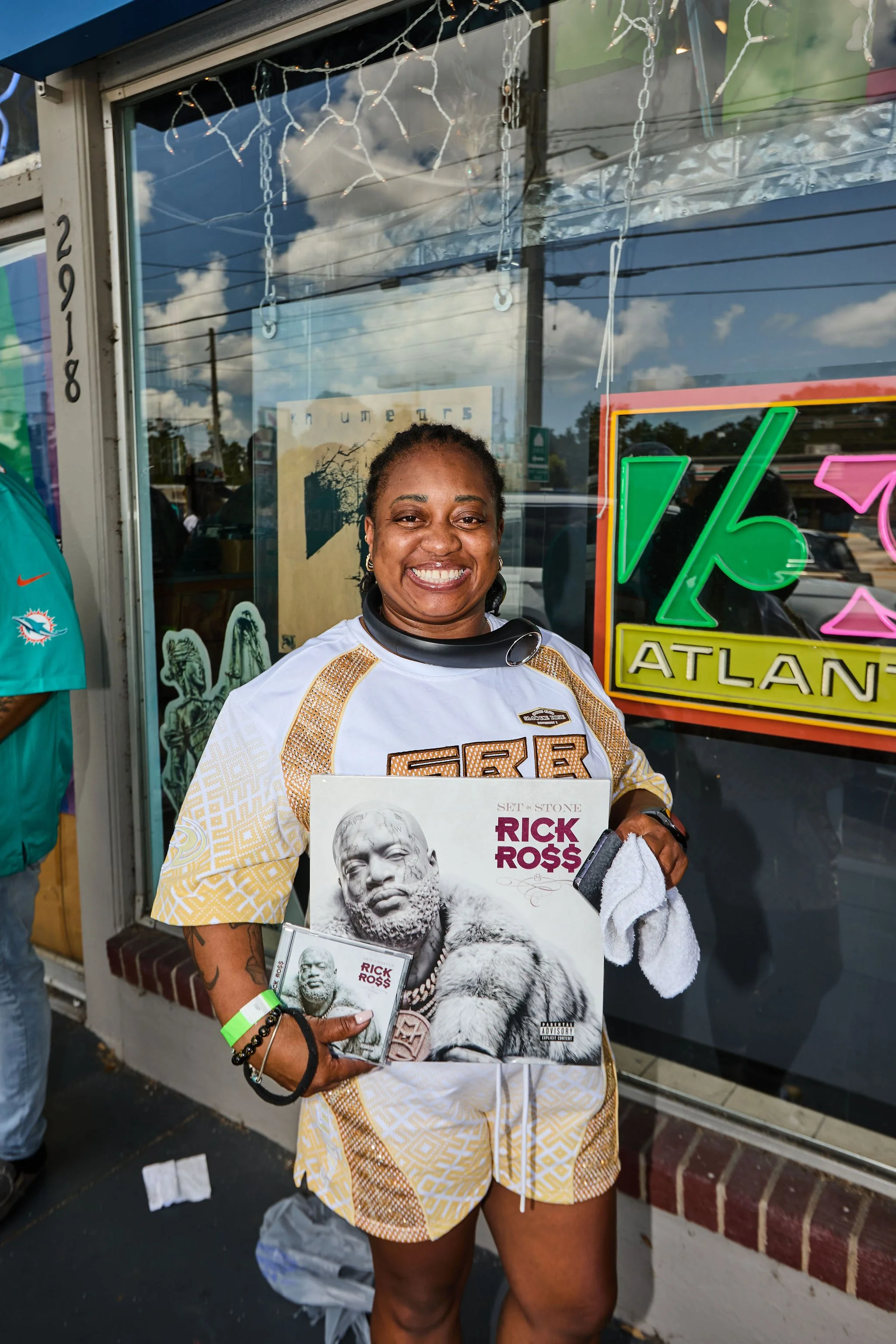 A woman smiling while holding a Rick Ross album in front of a storefront window with neon and string lights. She is wearing a white and gold sports jersey, and has tattoos on her arms. The background includes reflections of clouds and power lines.