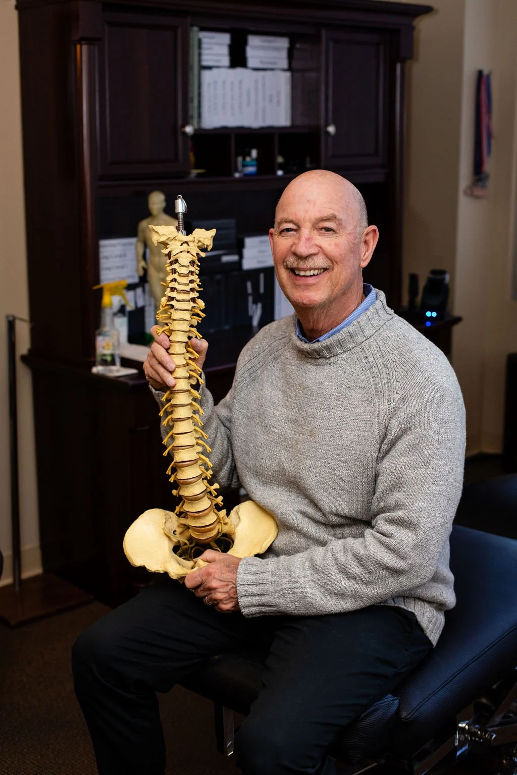 A smiling man in a gray sweater sitting in an office, holding a plastic spinal column model.
