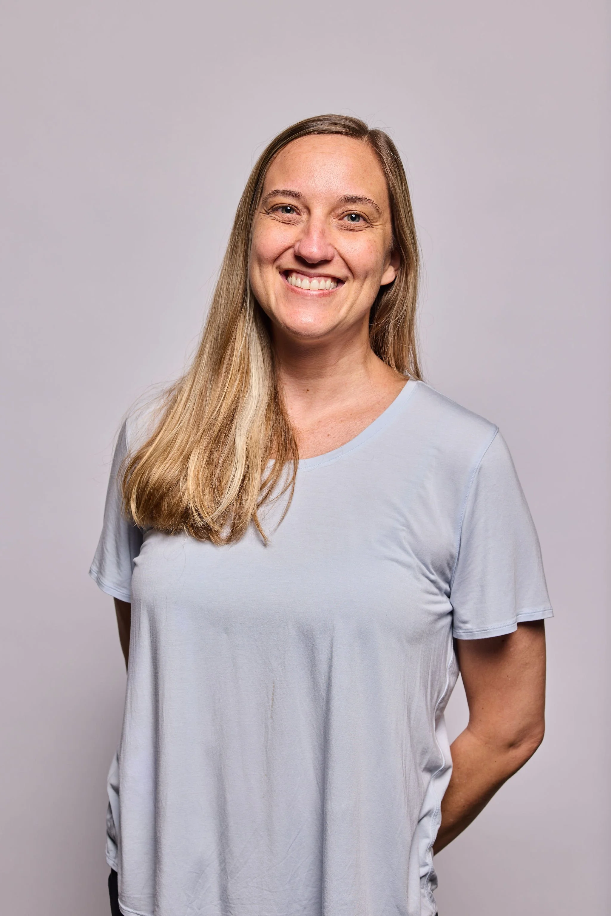 A smiling woman with long blonde hair wearing a light gray T-shirt, standing against a plain light gray background.