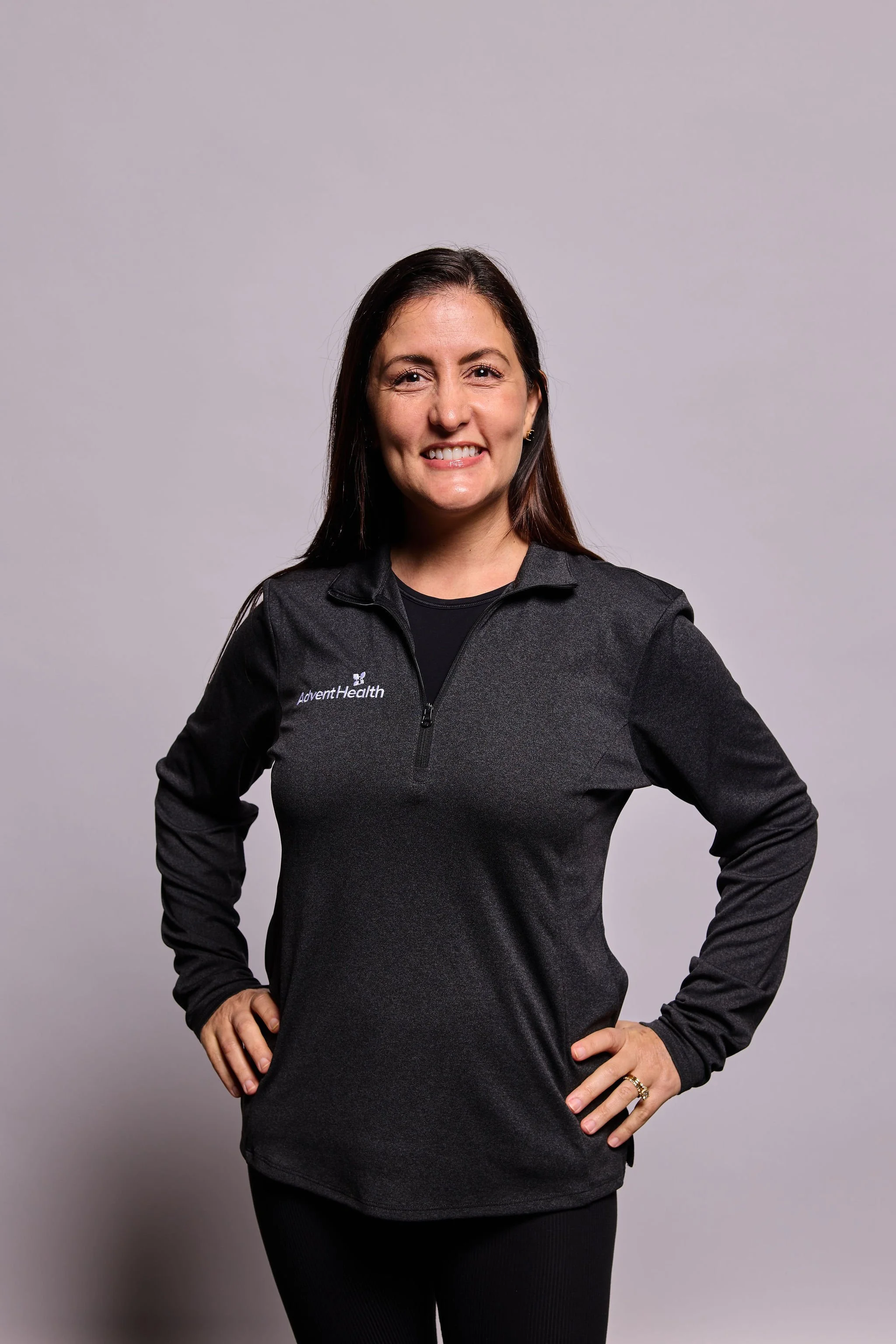 A woman with long dark hair, wearing a dark zip-up jacket with the text 'AdventHealth' on it, standing against a plain gray background, smiling, with one hand on her hip.