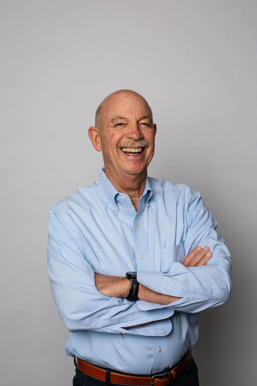 An older man with a bald head and a mustache, smiling and crossing his arms, wearing a light blue button-up shirt.