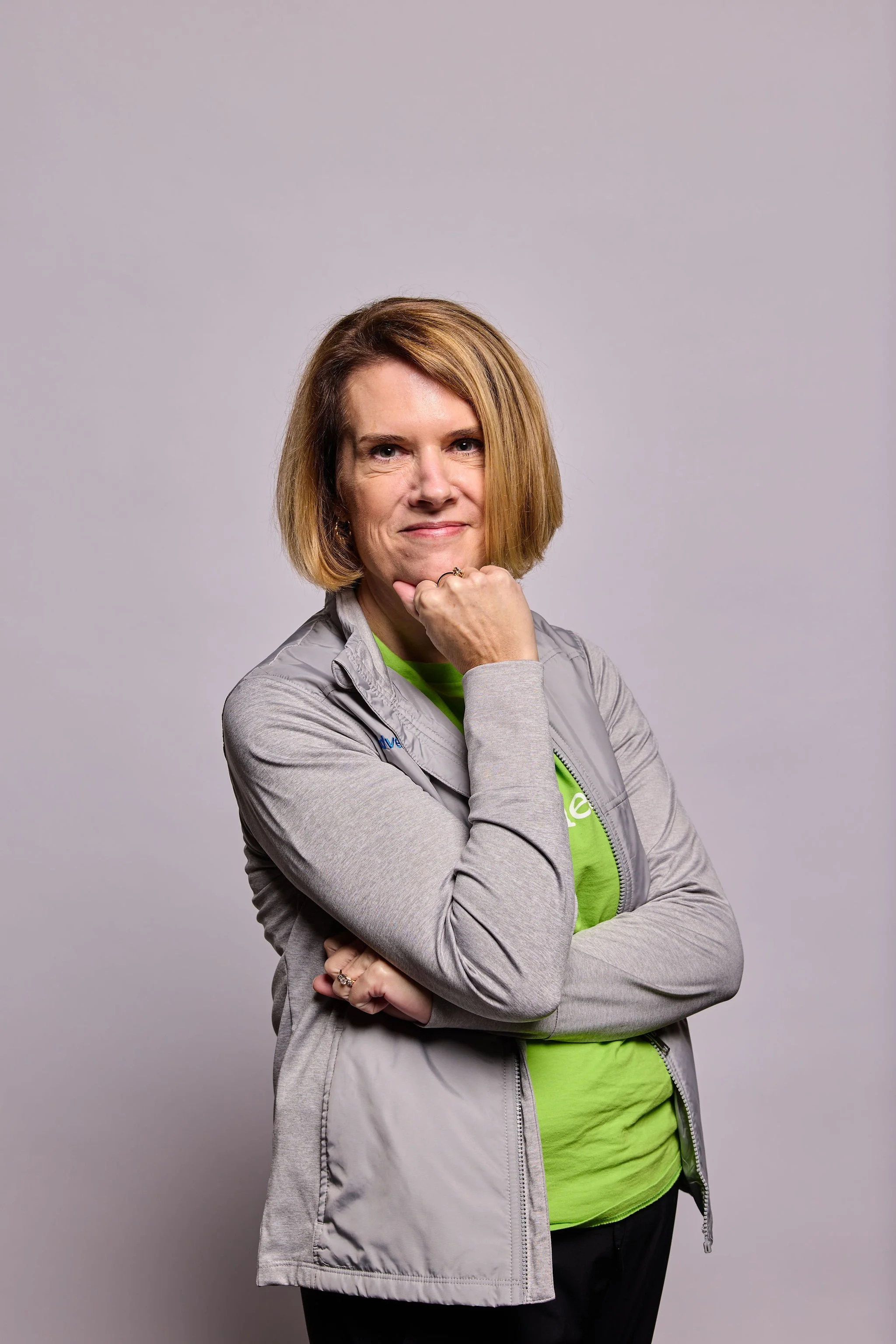 A woman with shoulder-length blonde hair wearing a gray jacket, neon green shirt, and black pants, standing with her arms crossed and resting her chin on her hand against a plain gray background.