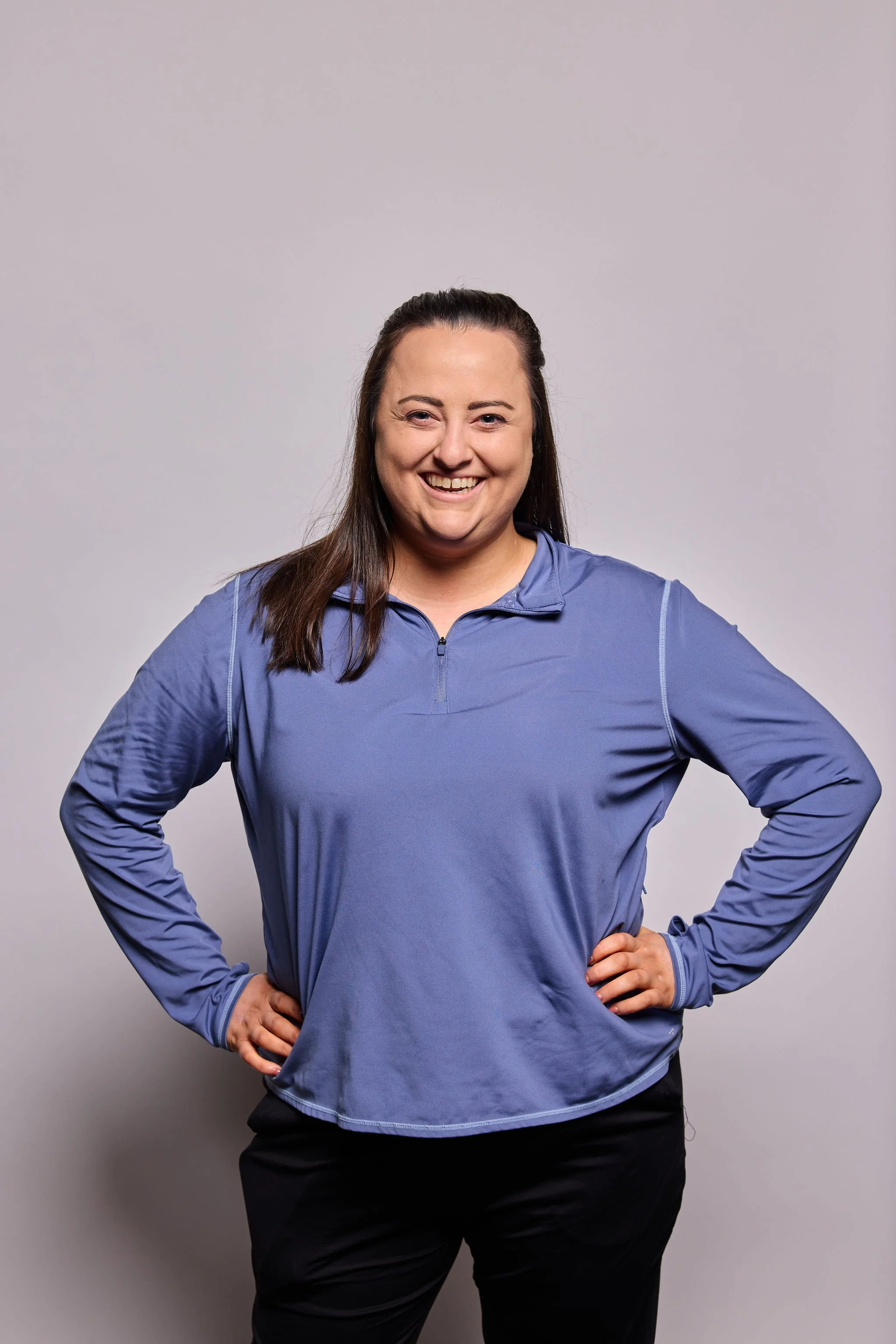 A woman with long brown hair wearing a blue long-sleeve athletic shirt and black pants, standing against a plain gray background, smiling with her hands on her hips.