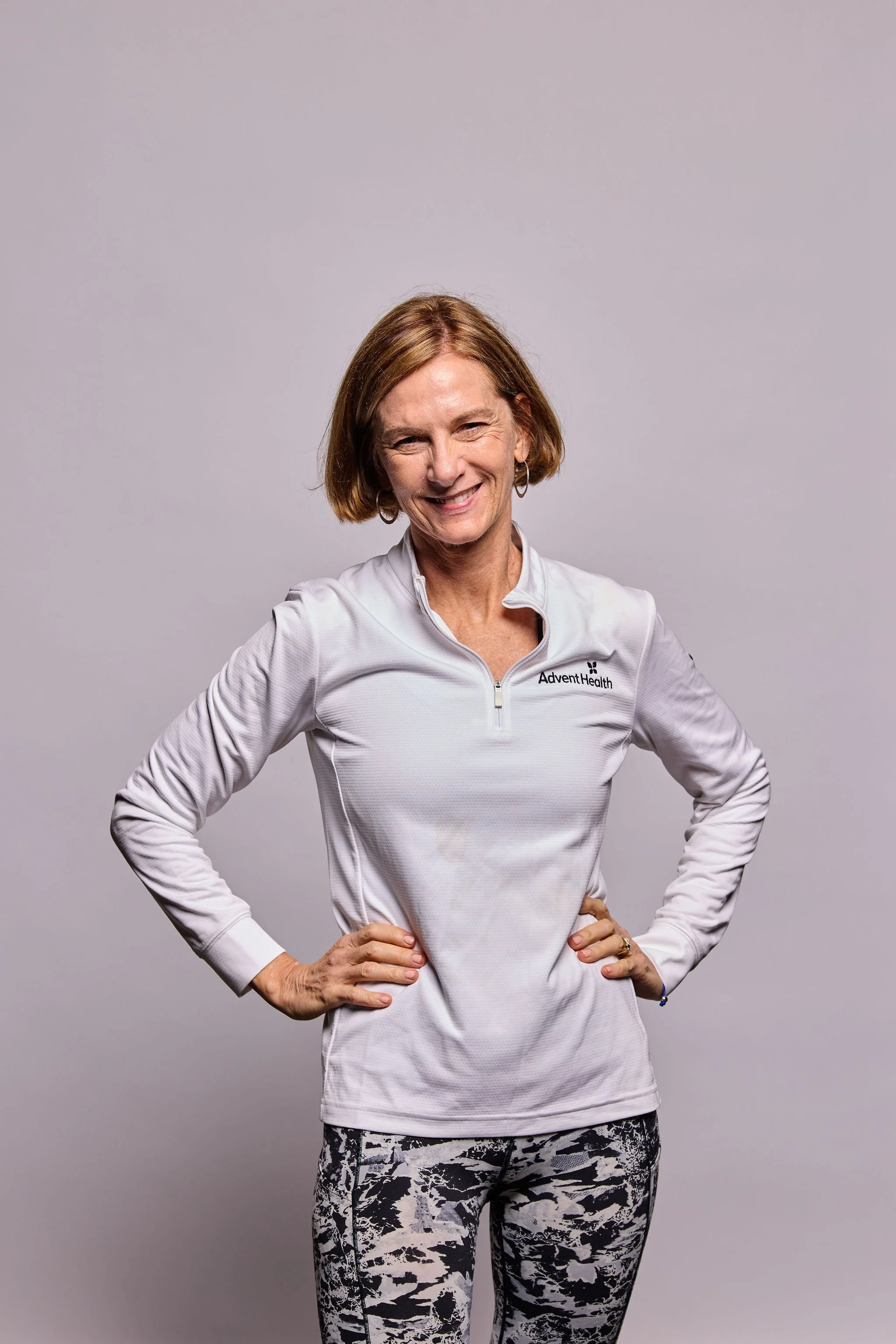 A middle-aged woman with short brown hair wearing a white athletic jacket with 'AdventHealth' logo and black and white patterned leggings, standing with hands on hips against a plain light gray background.