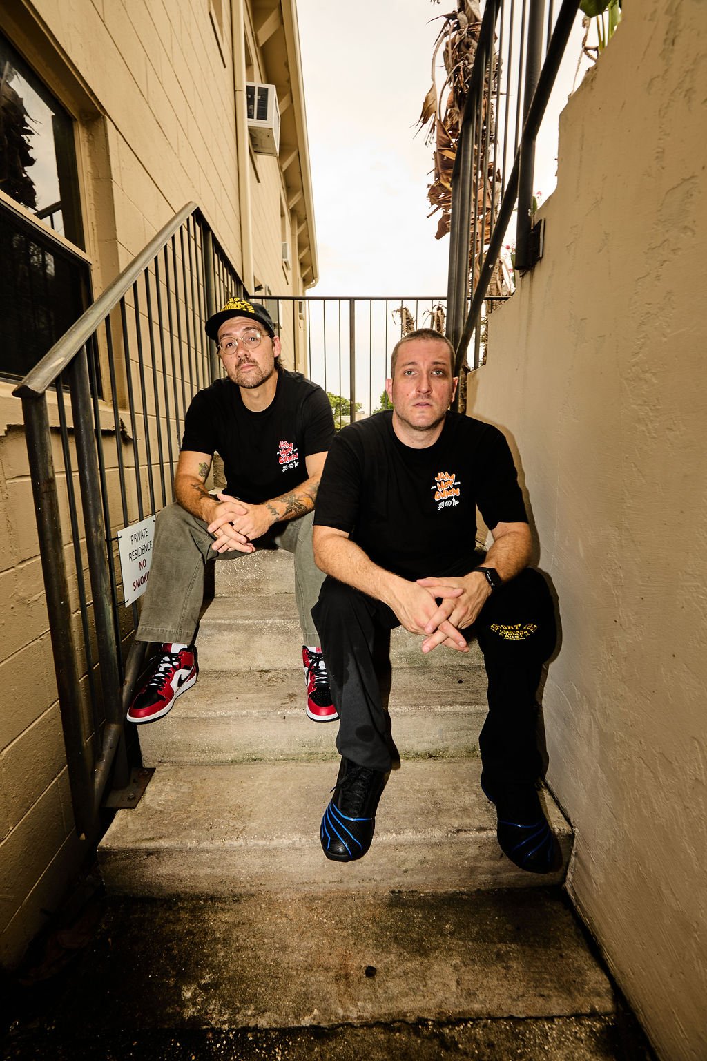 Two men sitting on outdoor stairs, wearing black shirts and sneakers, with beige walls and a railing behind them.