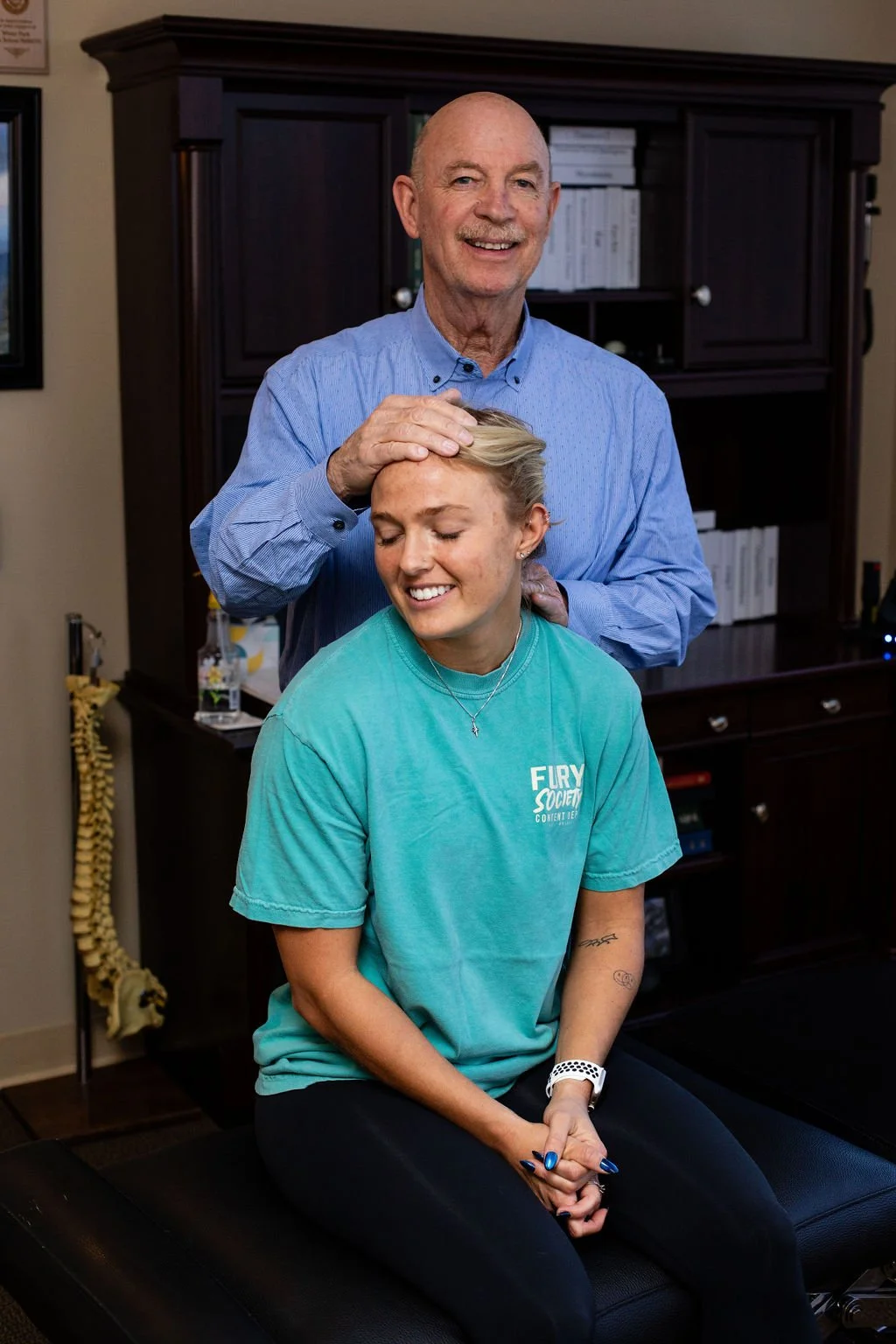 An elderly man in a blue shirt giving a chiropractic adjustment to a young woman in a turquoise t-shirt sitting on an examination table in a clinic. The man has a gentle smile and is placing his hand on the woman's head, who has her eyes closed and is smiling.