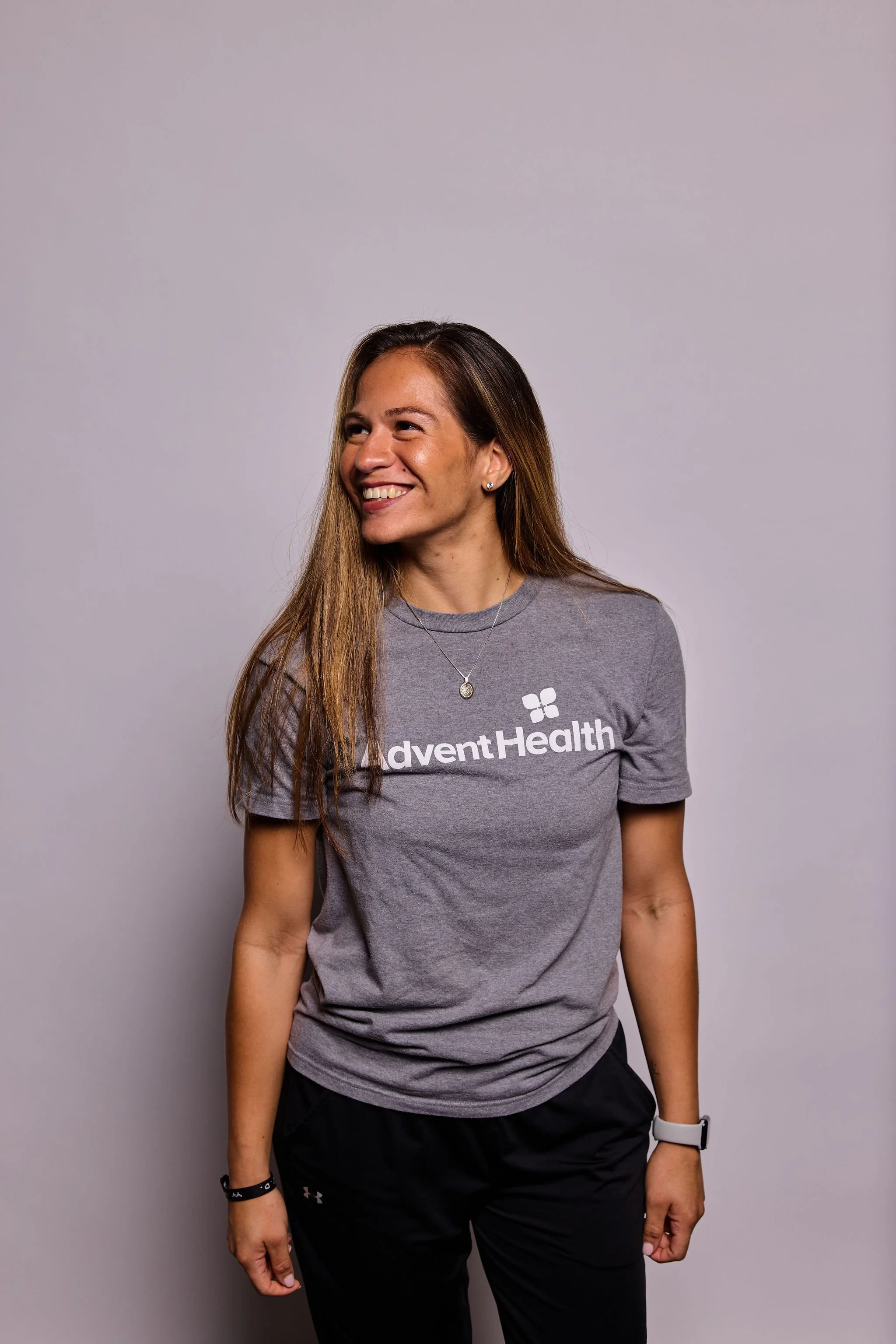 A woman in a gray t-shirt with 'adventHealth' logo, black pants, smiling and looking to her left, standing against a plain gray background.