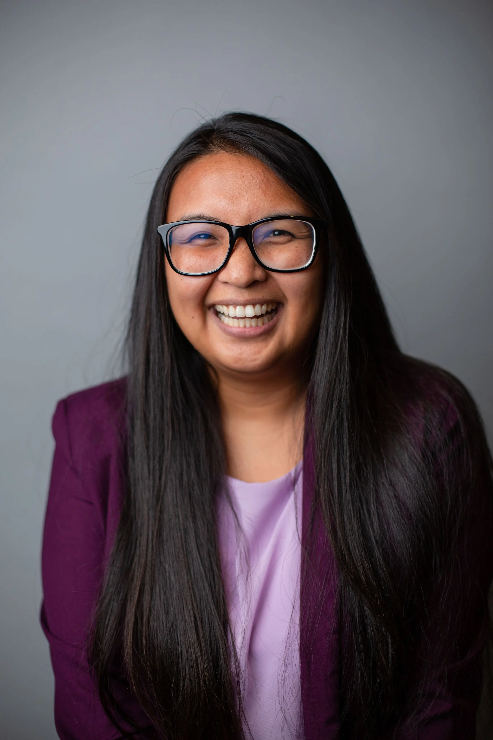 A woman with long dark hair, wearing glasses, a maroon blazer, and a light purple top, smiling in front of a plain gray background.