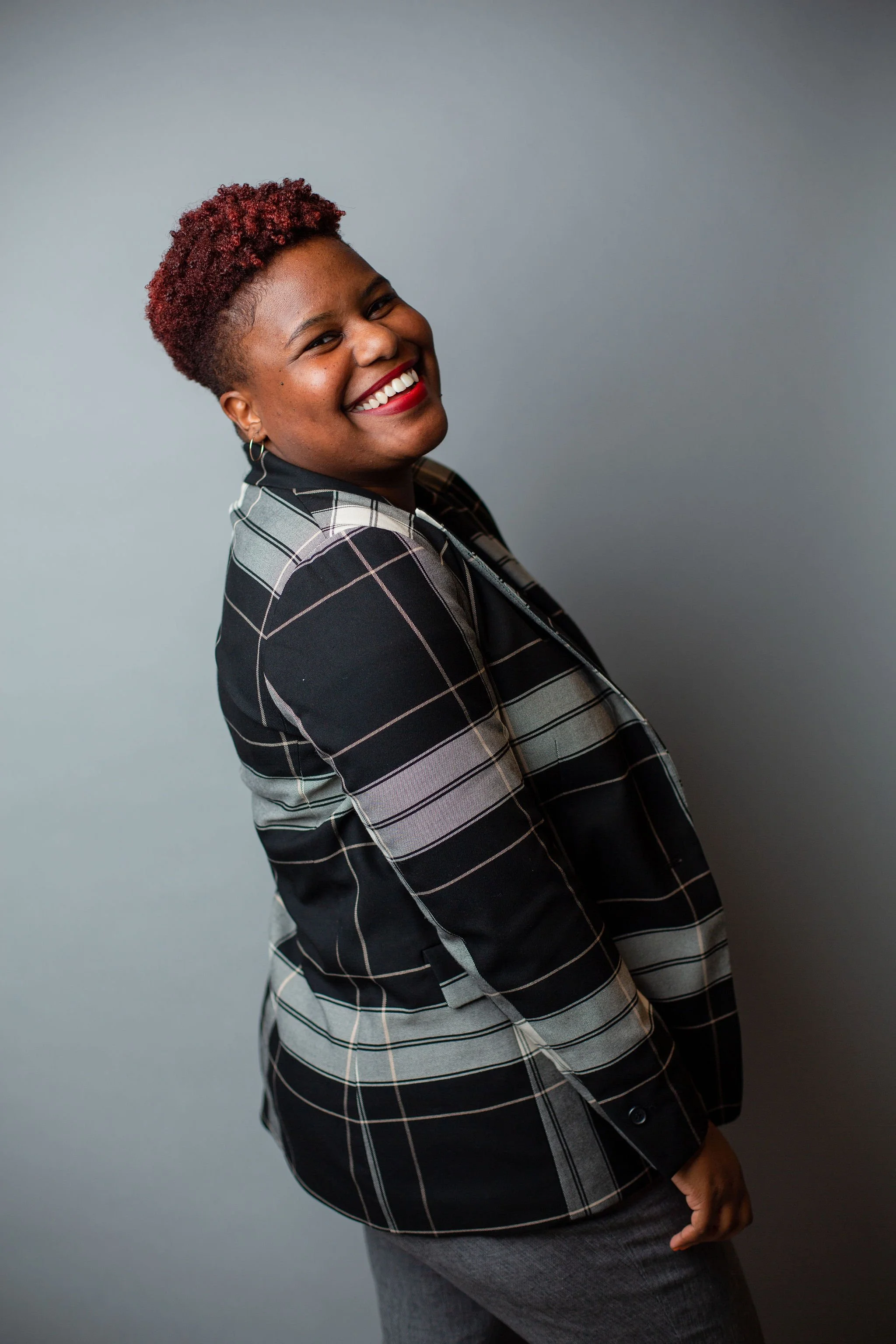 A woman with short, curly, reddish hair smiling and looking at the camera, wearing a black and white plaid blazer and gray pants, standing against a plain gray background.