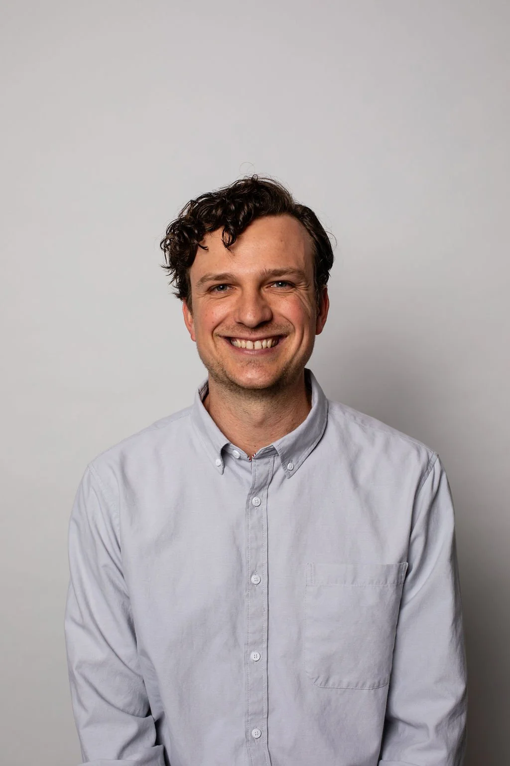A smiling man with dark, curly hair wearing a light gray button-up shirt standing against a plain light gray background.