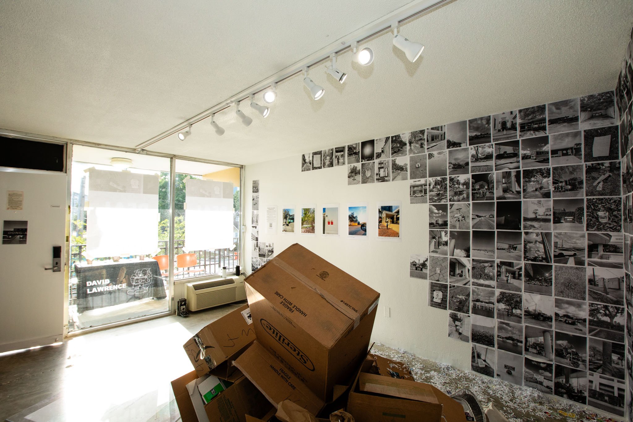 Empty art gallery with black and white photographs on the walls, some color photographs, cardboard boxes on the floor, and large front windows letting in sunlight.