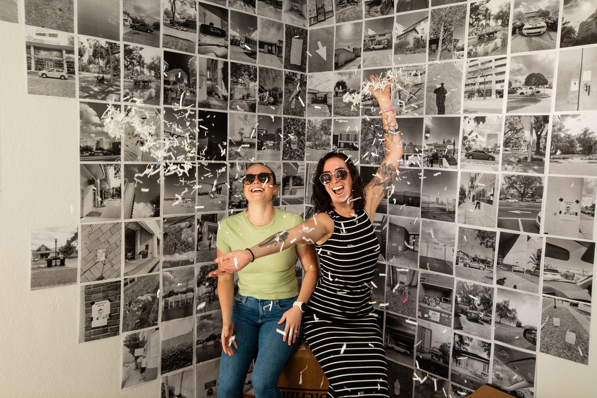 Two women celebrating with confetti in front of a wall featuring black and white photos of various buildings and street scenes.