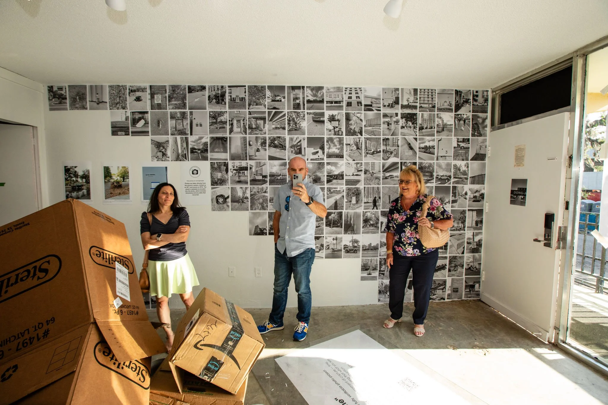 Three people standing inside an art gallery with black-and-white photos on the wall; a man is taking a selfie, another woman is smiling, and a third woman is standing with arms crossed; cardboard boxes are on the floor near the window.