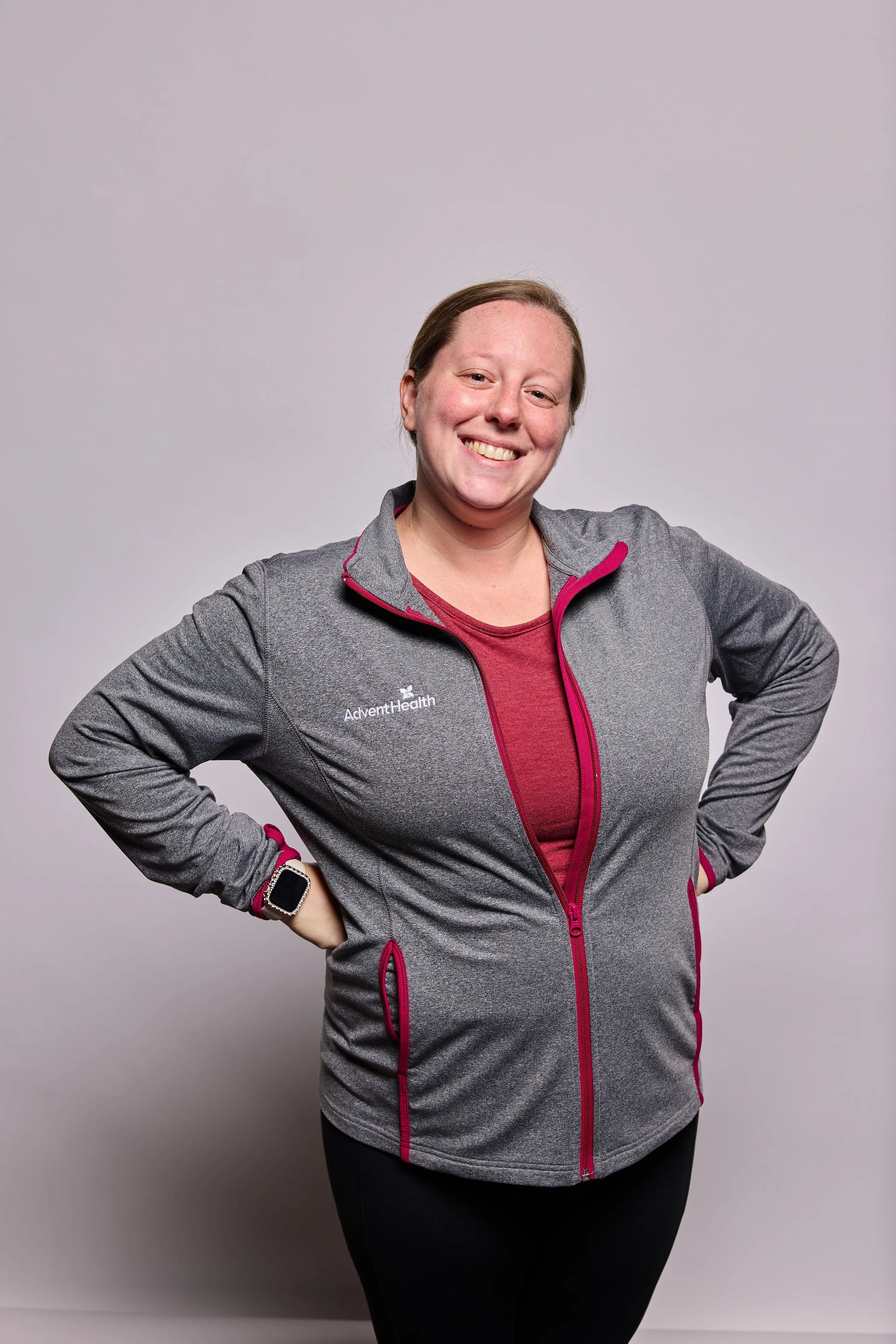 A smiling woman with light brown hair wearing a gray and magenta athletic jacket with 'AdventHealth' logo, black leggings, a smartwatch, standing against a plain light gray background.