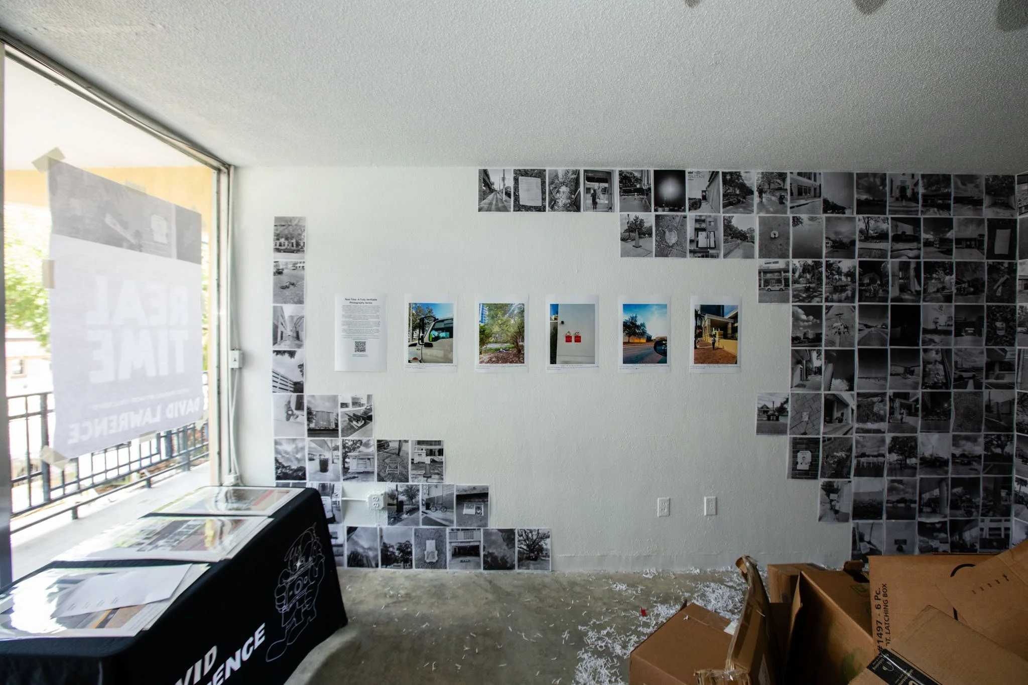 Interior of an art gallery with a wall featuring collage of black-and-white photographs and color photographs attached in the middle. A large window on the left side has a sign taped to it. There are boxes on the floor in the foreground.