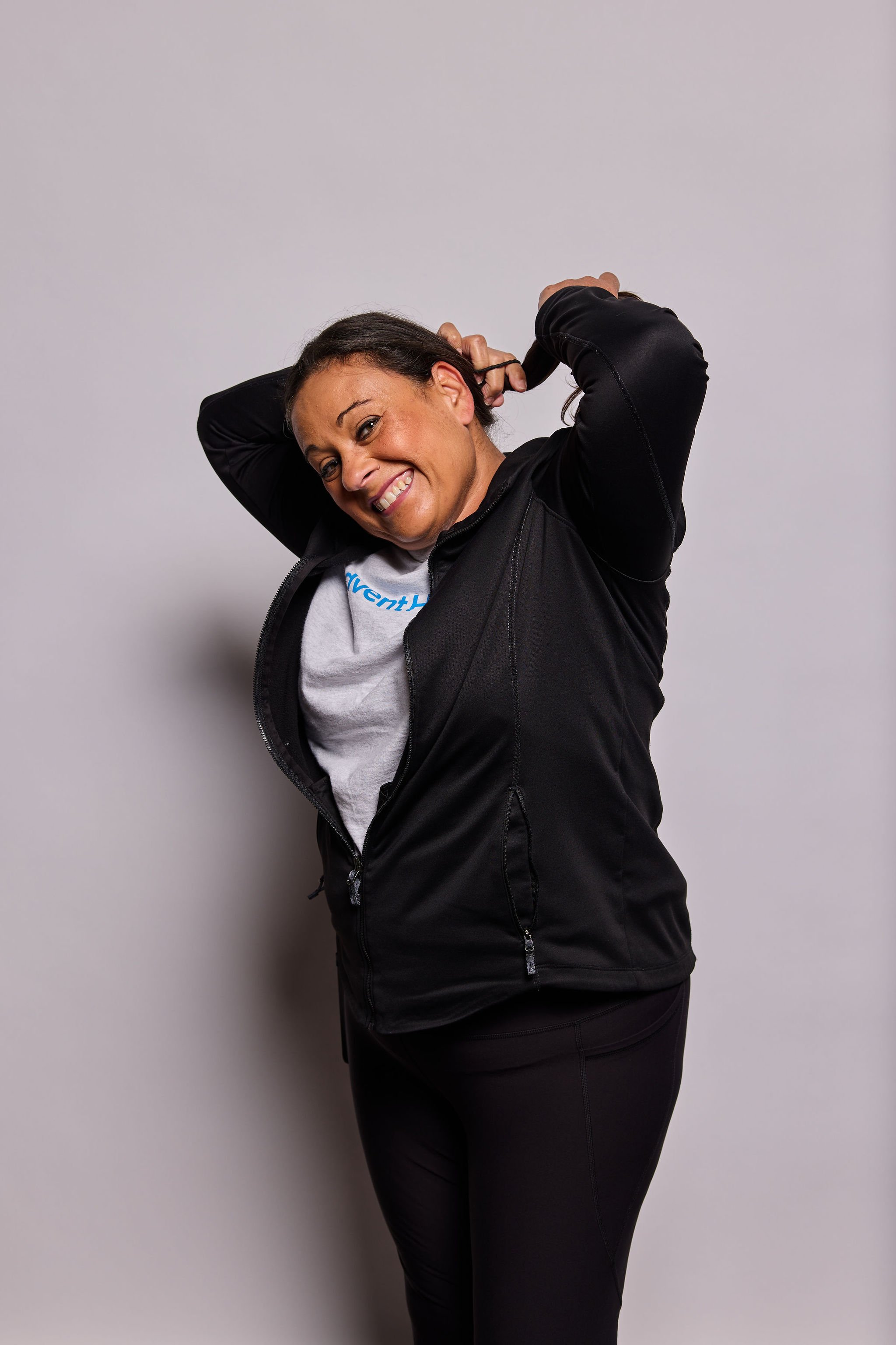 Smiling woman in black jacket and black pants standing against a plain gray background, with her arms raised behind her head.