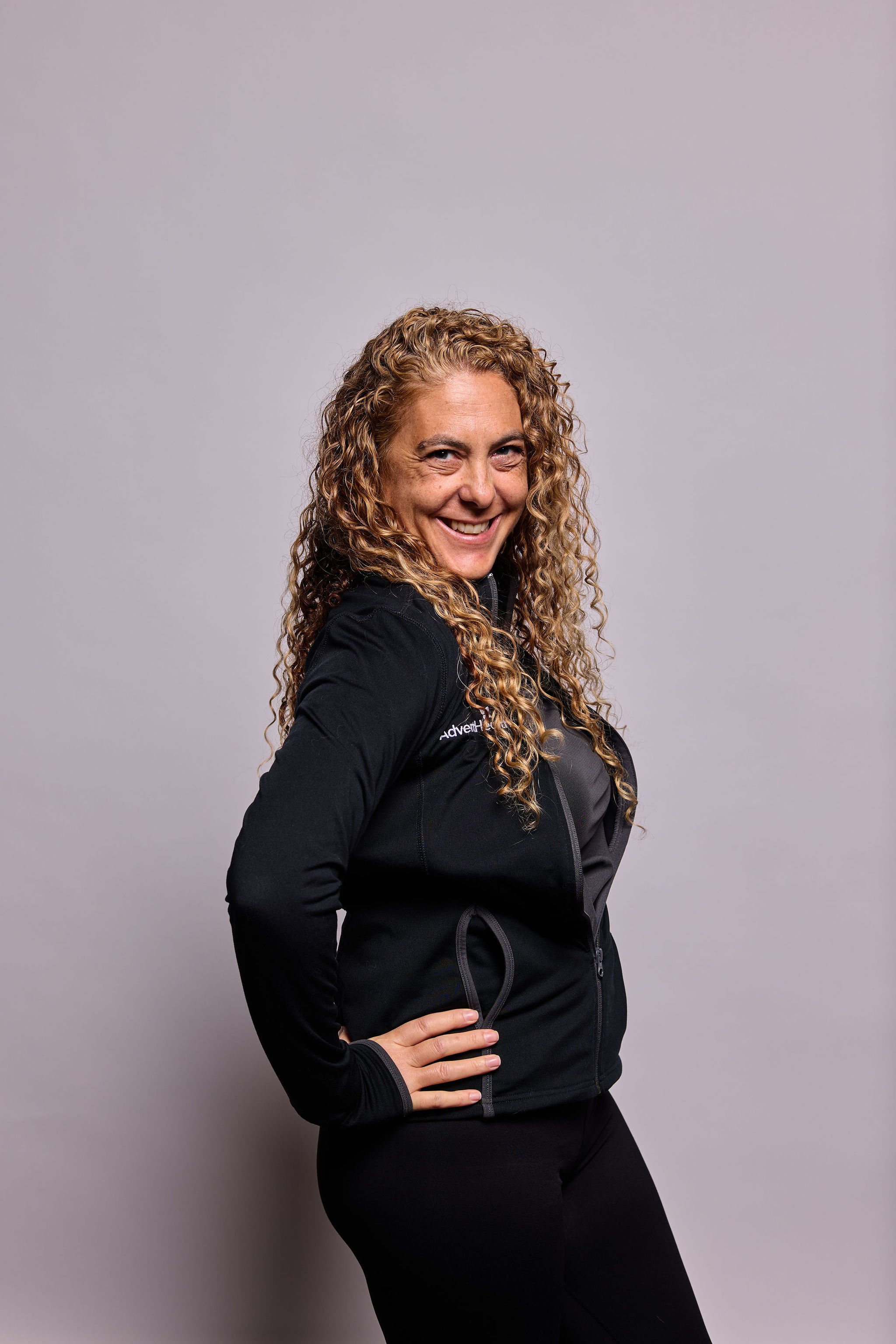 A woman with curly blonde hair smiling, standing with her hand on her hip, wearing a black athletic jacket against a light gray background.