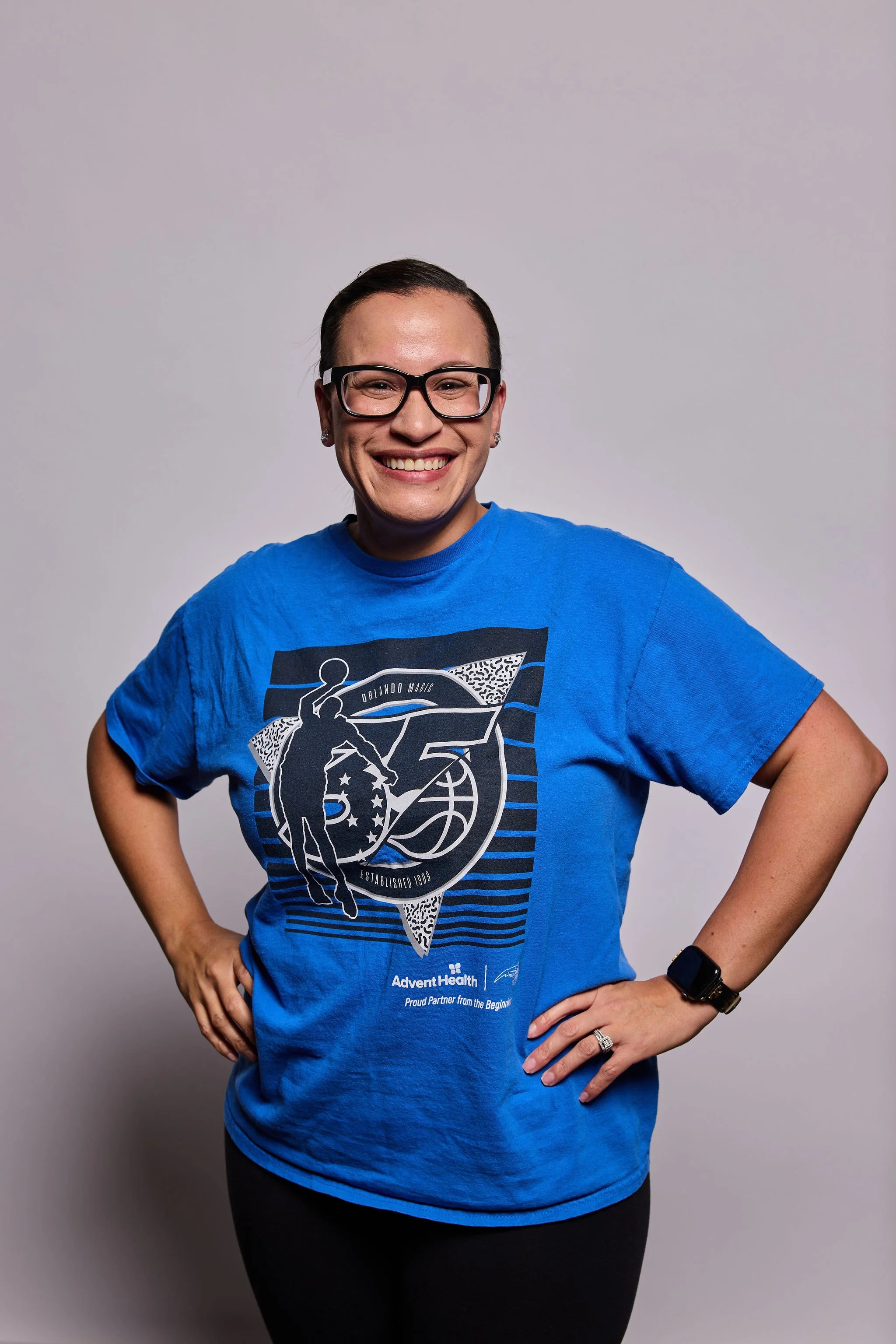 A woman with short dark hair, glasses, and a big smile wearing a blue Orlando Magic T-shirt and black leggings standing against a plain wall.