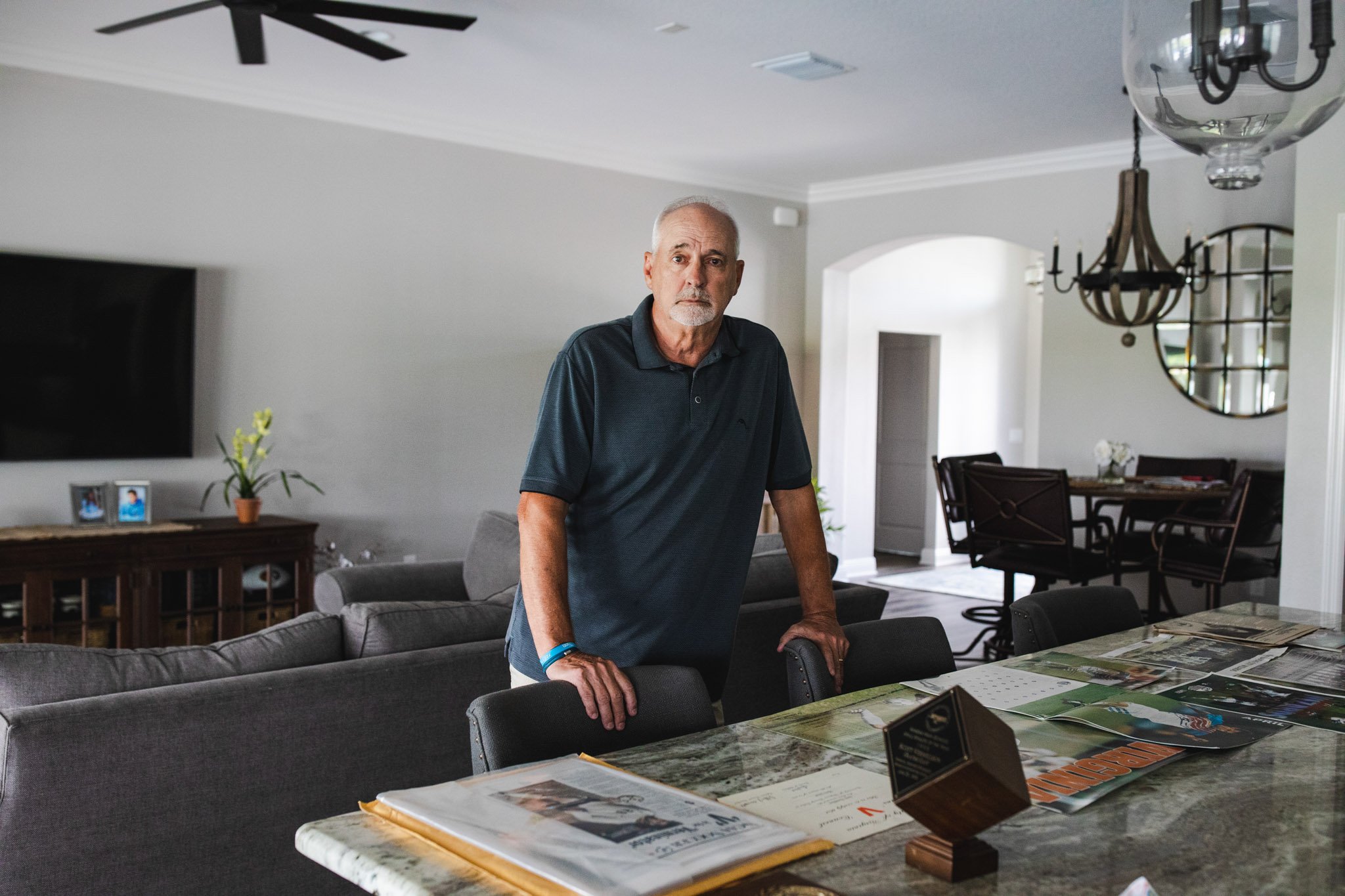 An older man with gray hair and a beard stands behind a dining table in a modern, well-lit living room, looking at the camera.