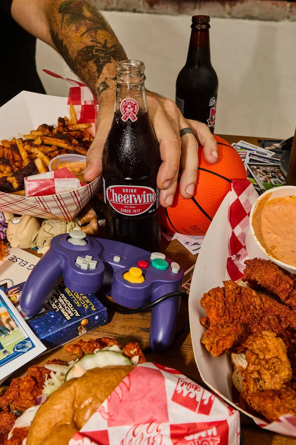Person with tattoos holding a Cheerwine soda bottle, surrounded by fried chicken, fries, a basketball, a gaming controller, and various food items on a table.