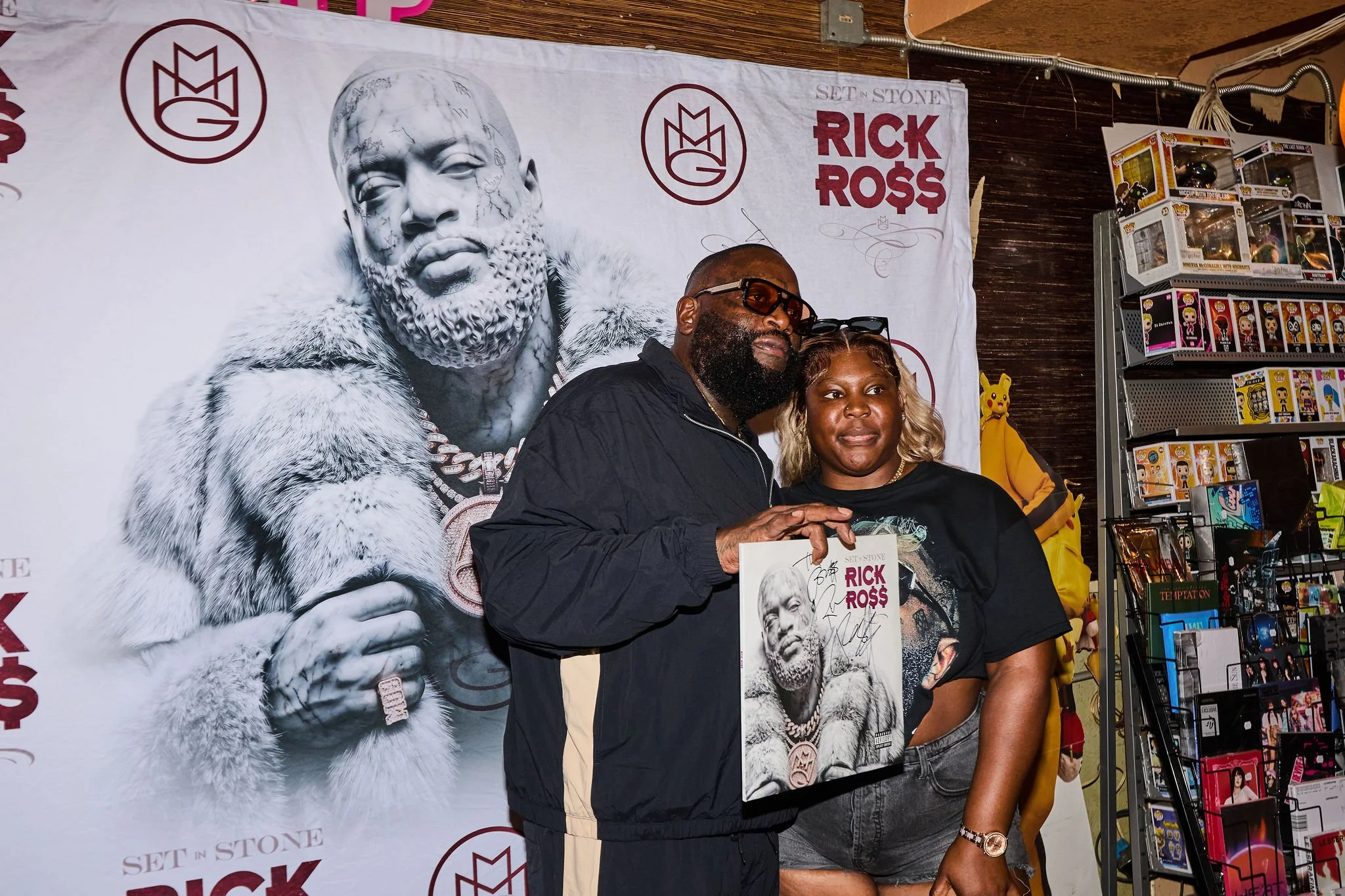 Rick Ross at a signing event holding an autographed photo of himself with a large poster of himself in the background and shelves of collectibles nearby.