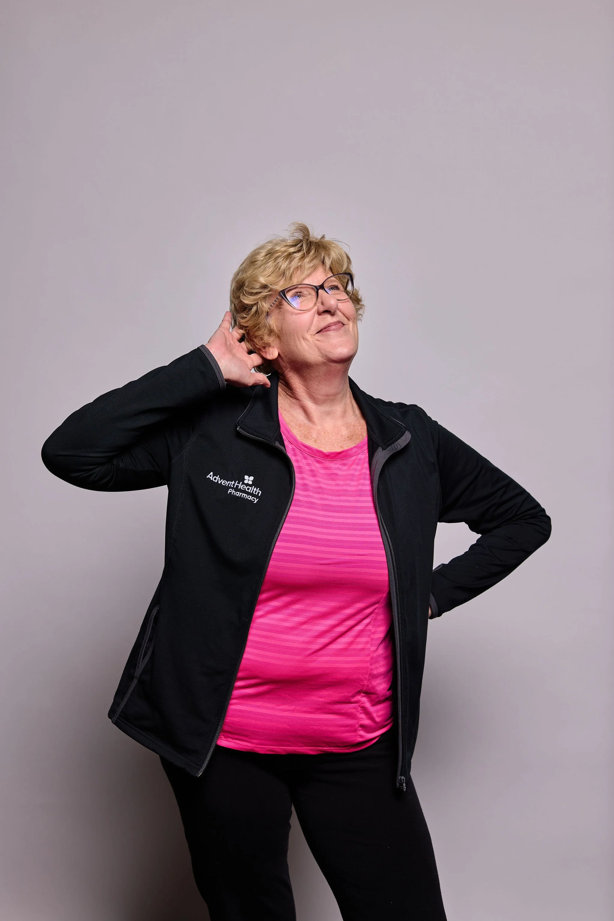 A woman with short blonde hair, glasses, and a black jacket with 'AdventHealth Pharmacy' logo, standing against a light gray background, posing with a smile, one hand behind her head, and wearing a bright pink shirt.