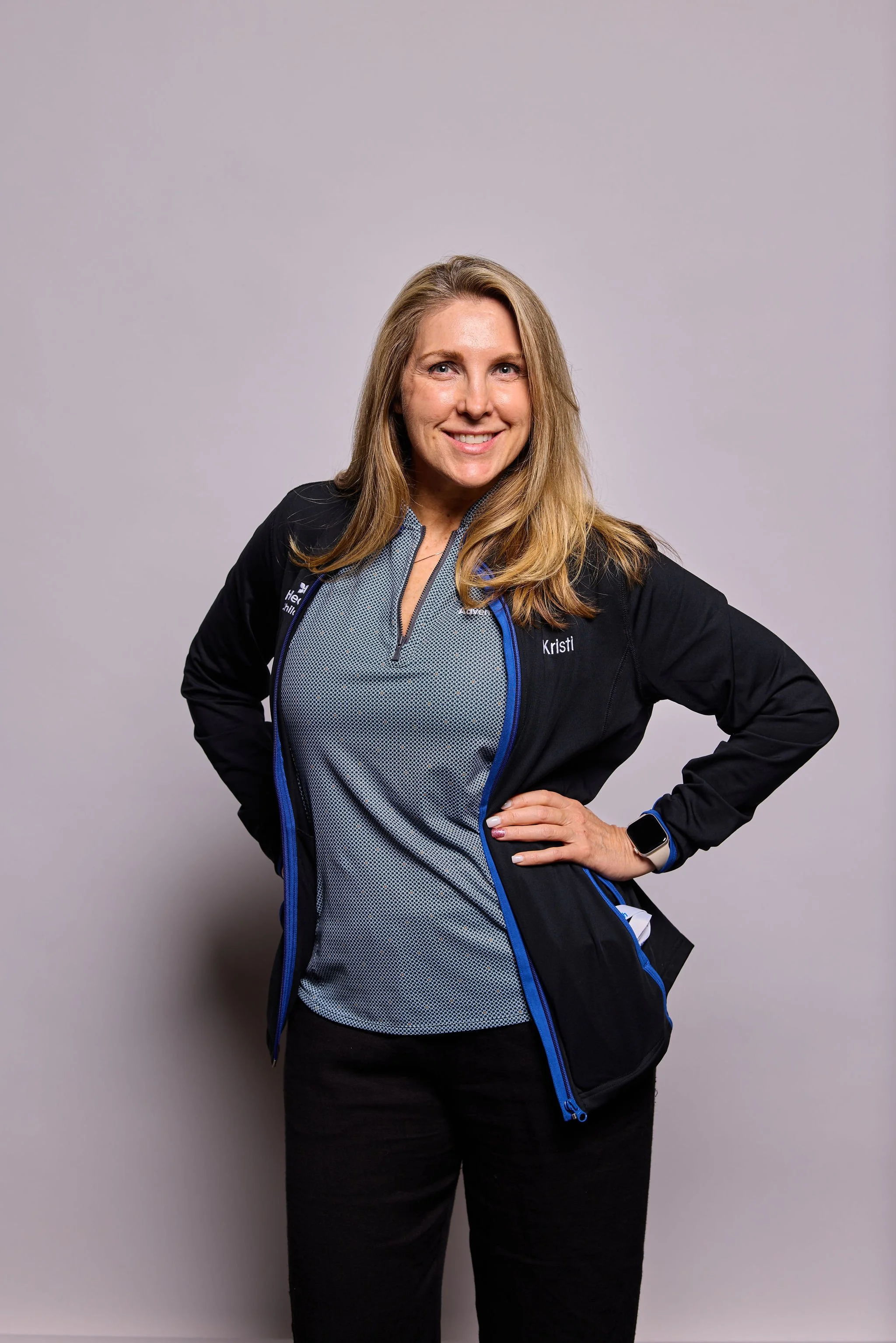 A woman with long blonde hair, wearing a black jacket with blue accents and a gray athletic top, smiling and posing with her right hand on her hip against a plain gray background.