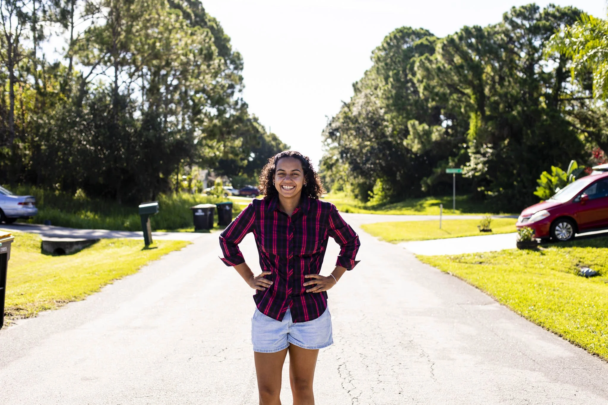 A smiling young woman with curly hair standing in the middle of a suburban street with her hands on her hips, wearing a red and black plaid shirt and denim shorts, with trees, cars, and a sunny sky in the background.