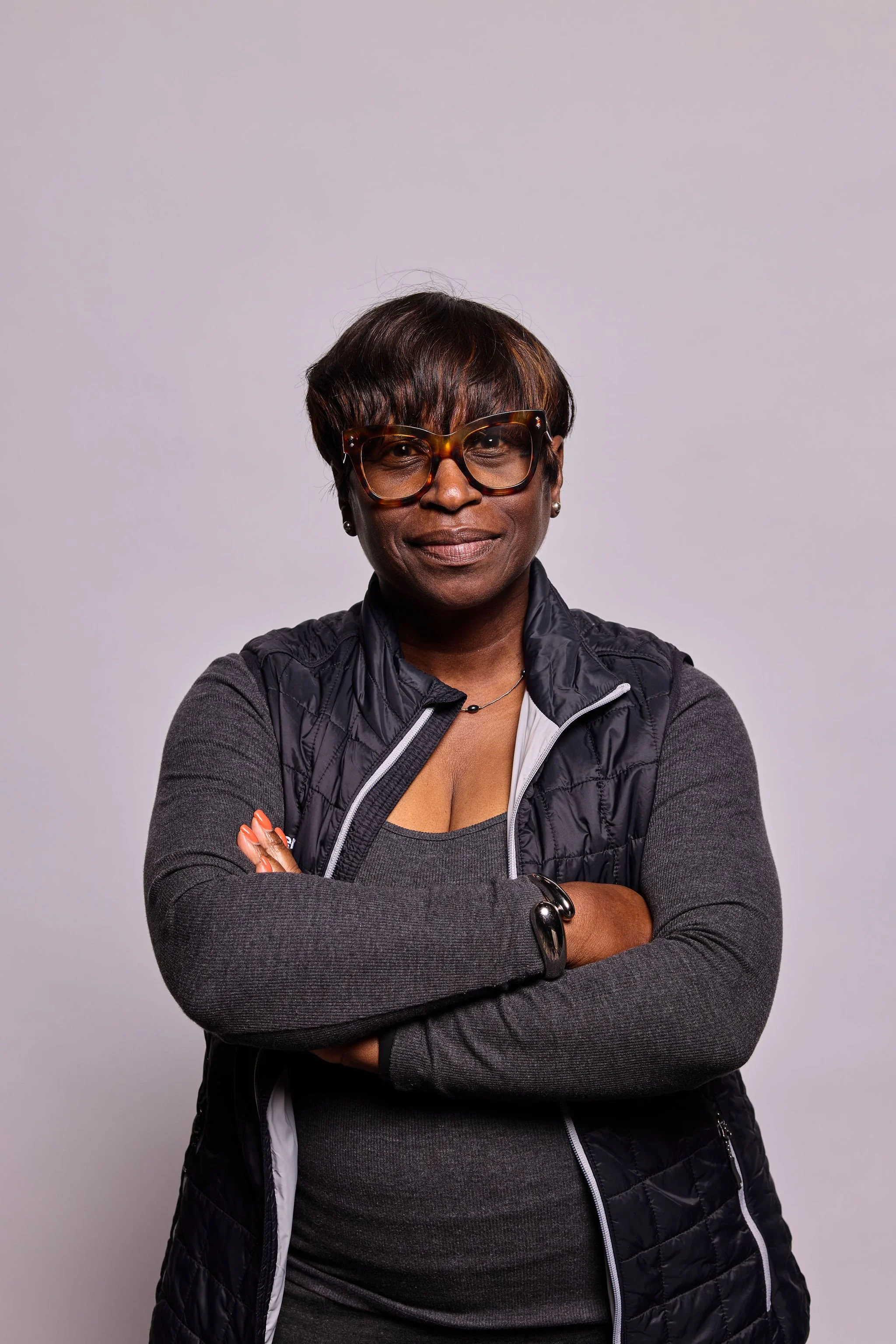 A woman with short dark hair wearing large tortoiseshell glasses, a black puffy vest over a gray long-sleeve shirt, and a silver bracelet, standing with arms crossed against a plain light gray background.