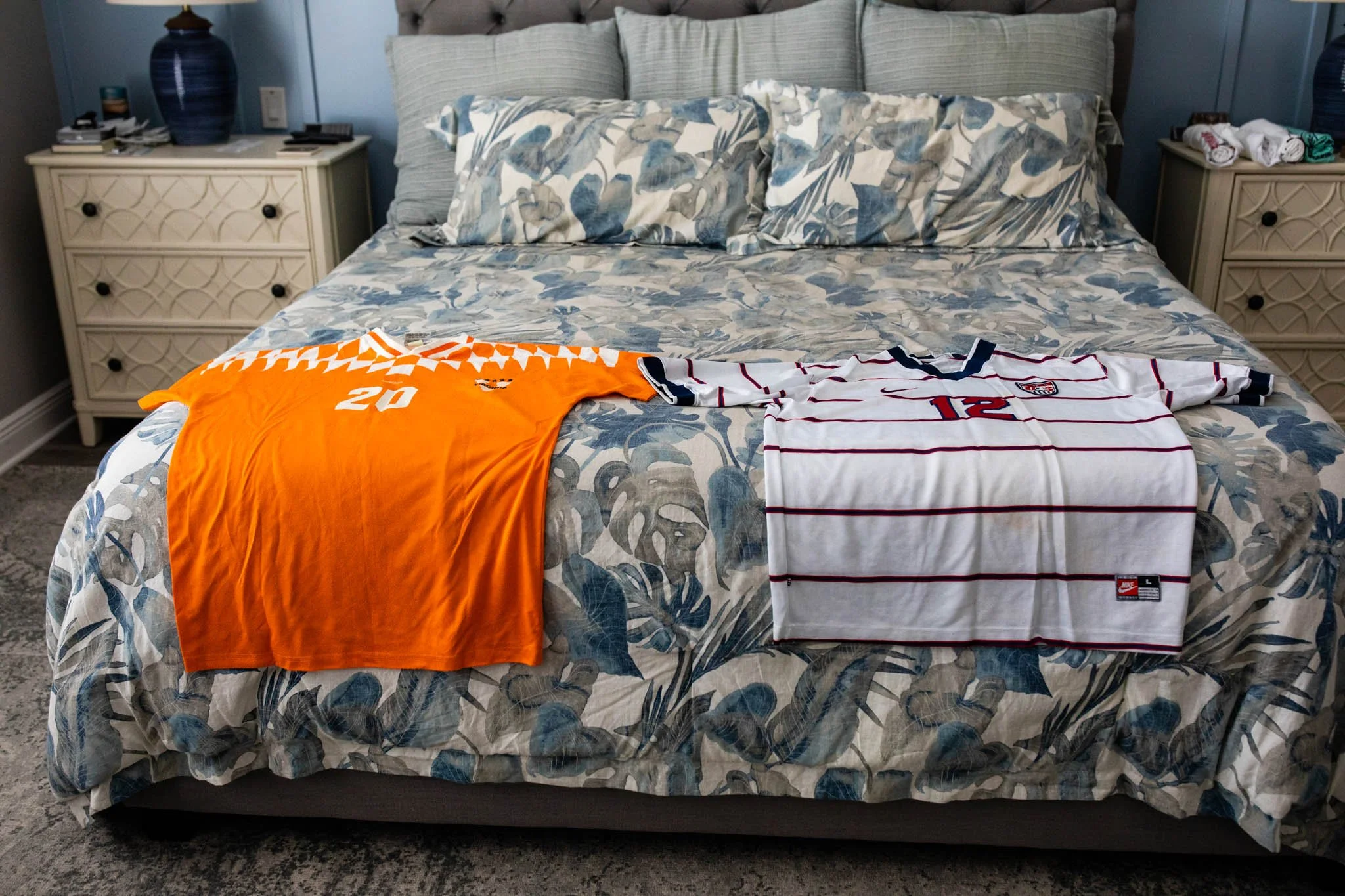 A bed with two sports jerseys laid out on top, one orange with the number 20, and one white with red and navy stripes with the number 12.