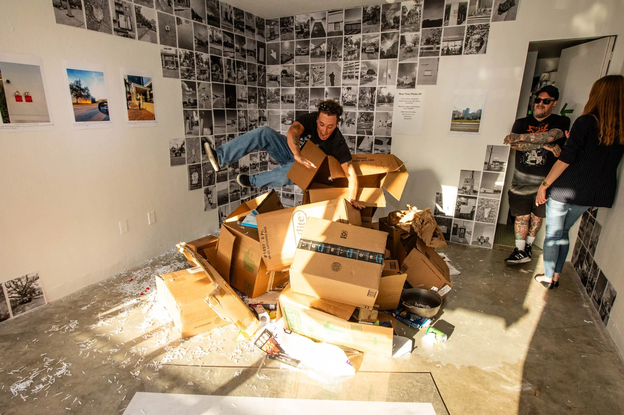 A person tumbling into a pile of cardboard boxes with a messy room in the background, while two other people stand and chat nearby.