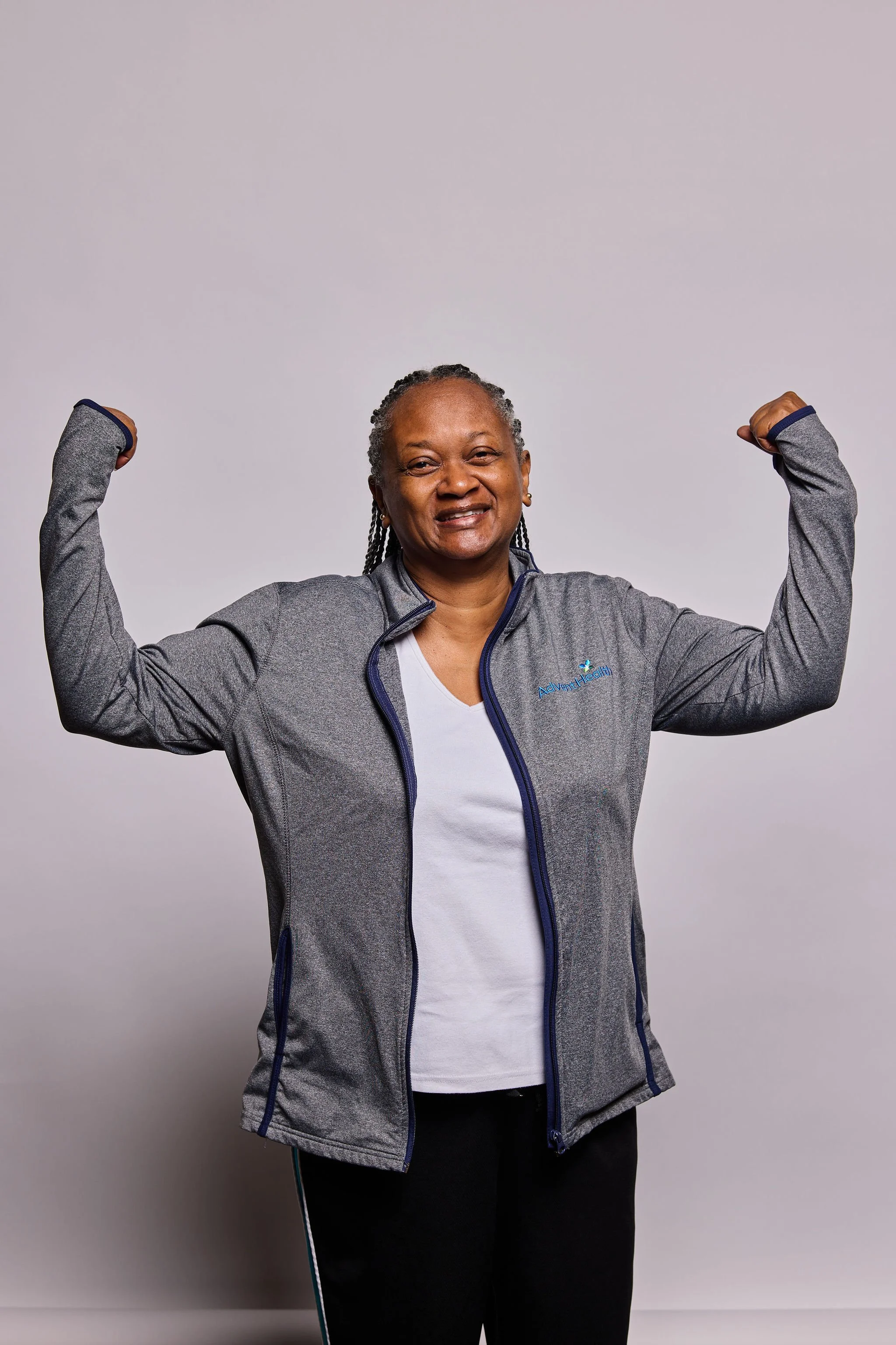 A smiling woman with gray braids flexing her biceps in front of a plain light-colored background. She wears a gray zip-up athletic jacket and black pants.