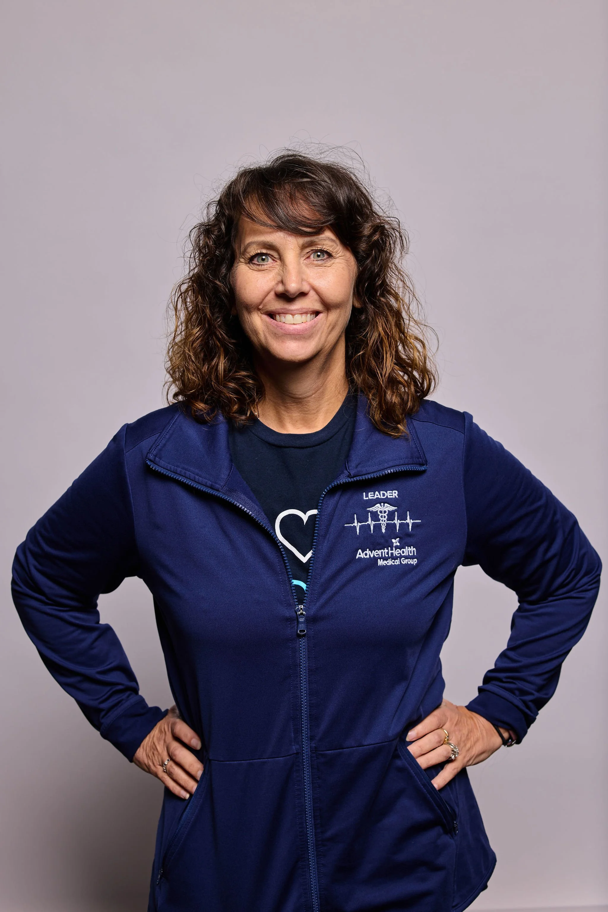 A woman with curly brown hair smiling, wearing a navy blue jacket with a Marine Corps Heartbeat logo and the words 'LEADER' and 'AdventHealth Medical Group' on it, standing against a plain gray background.