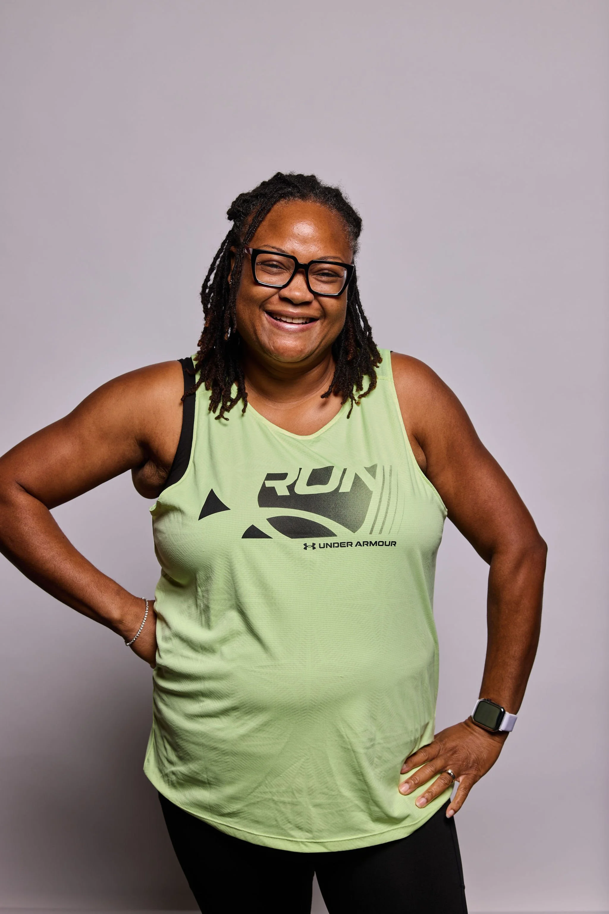 A smiling woman with glasses, dreadlocks, wearing a bright yellow Under Armour tank top and black leggings, standing against a plain gray background with her hands on her hips.