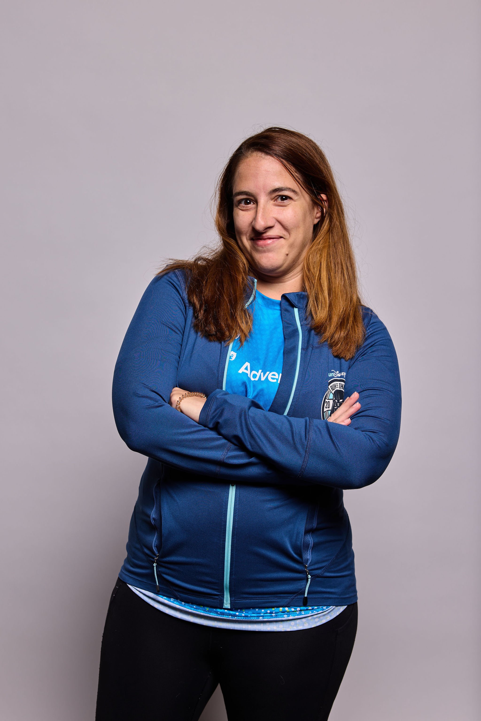 A woman with long brown hair wearing a blue jacket with various patches and a Nike logo, standing against a plain gray background, with arms crossed and smiling slightly.