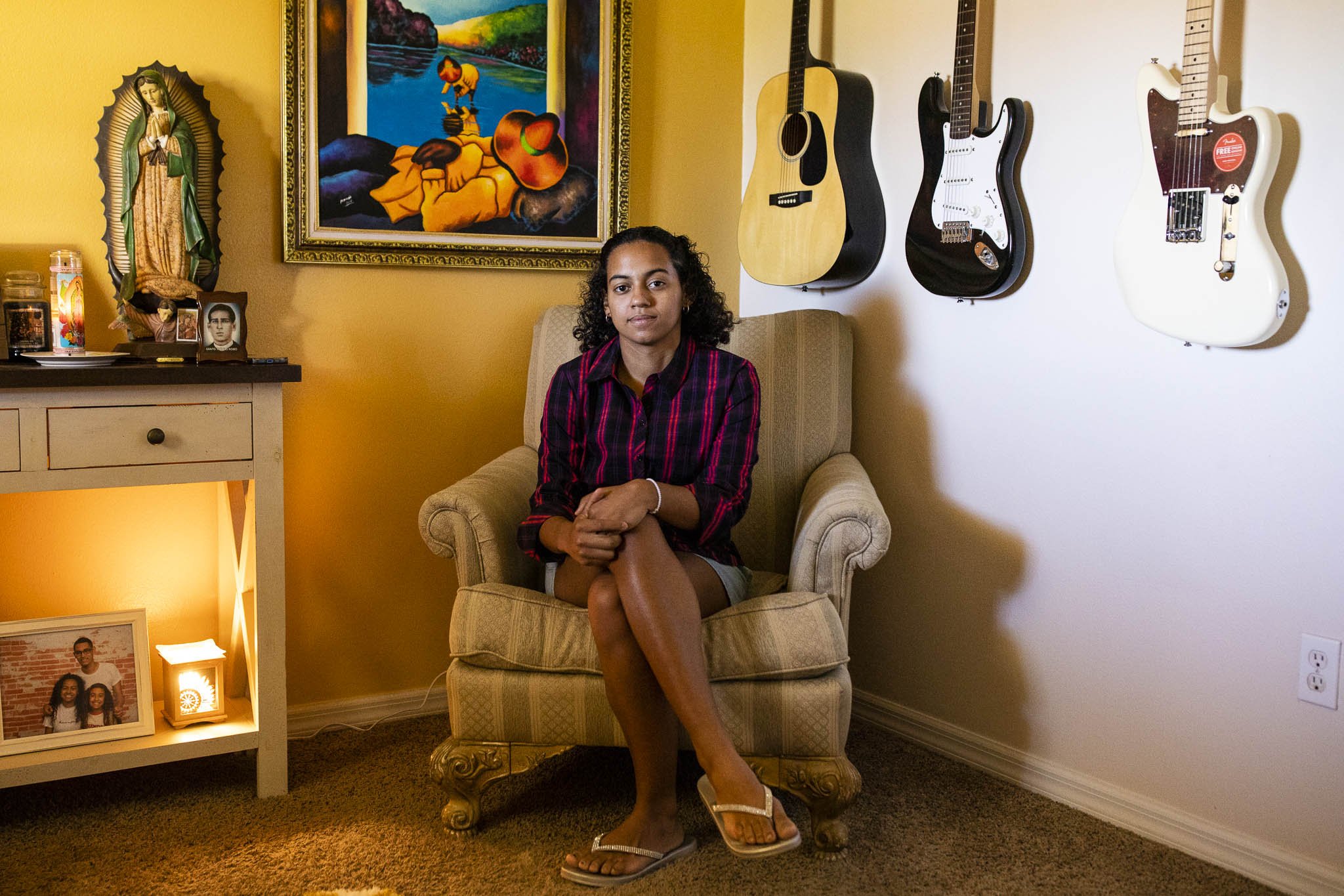 A young woman sitting in a beige armchair in a room with yellow walls, decorated with guitars, paintings, a religious statue, and framed photographs.