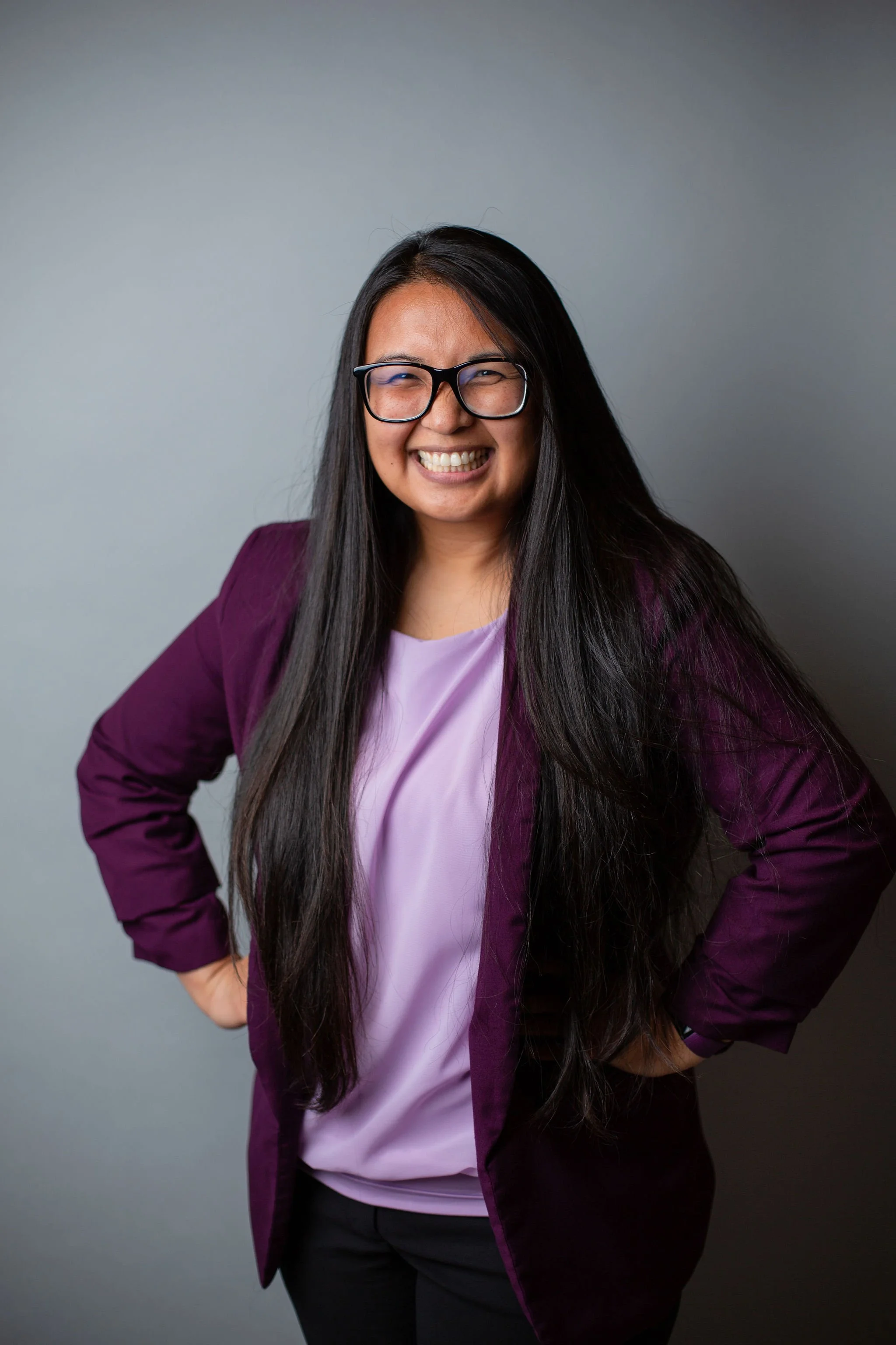 A woman with long black hair smiling, wearing glasses, a purple blazer, and a light purple blouse, standing against a gray background.