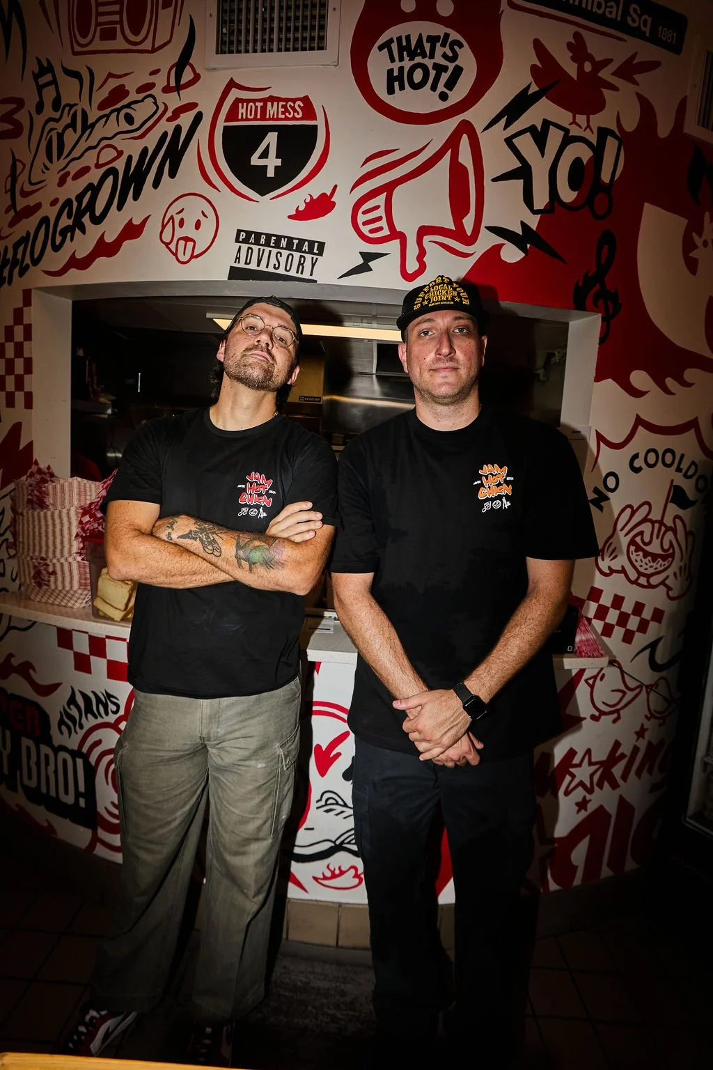 Two men stand in front of a graffiti-covered wall with various phrases and illustrations, wearing black t-shirts with logo designs, one with glasses and tattoos, the other with a cap.
