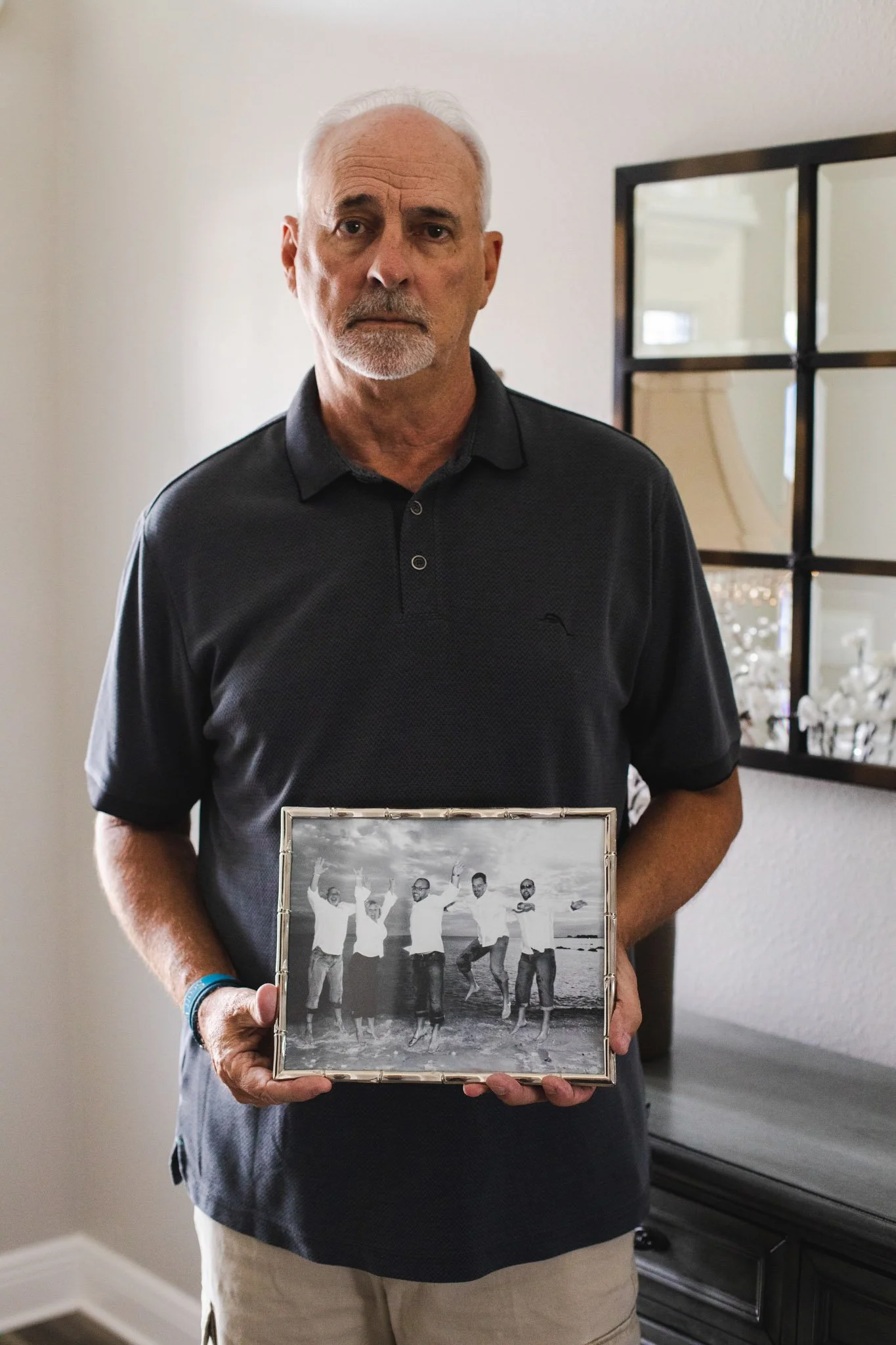 An older man with gray hair and a beard holding a framed black-and-white photo of five people at the beach, with the ocean and sky in the background.