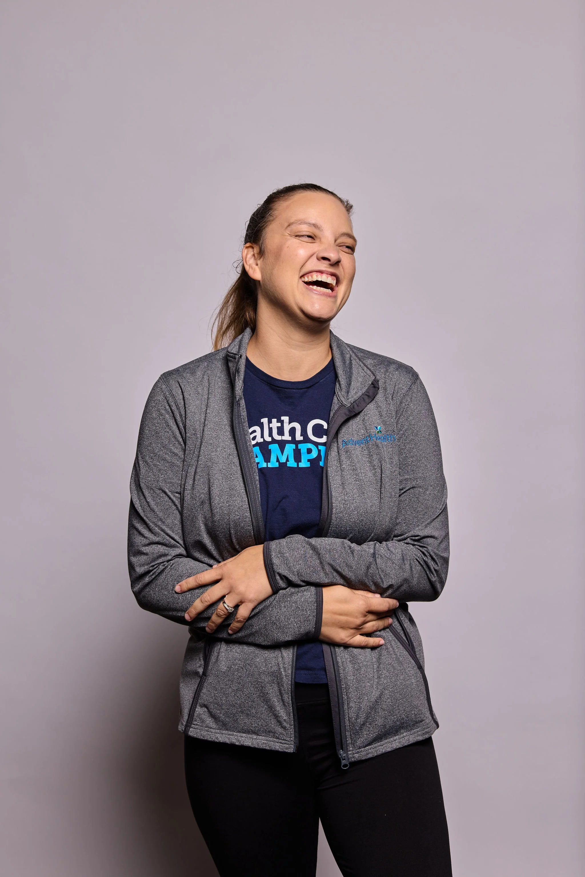A woman laughing, wearing a gray jacket with a logo on the chest, and black pants, standing against a plain gray background.
