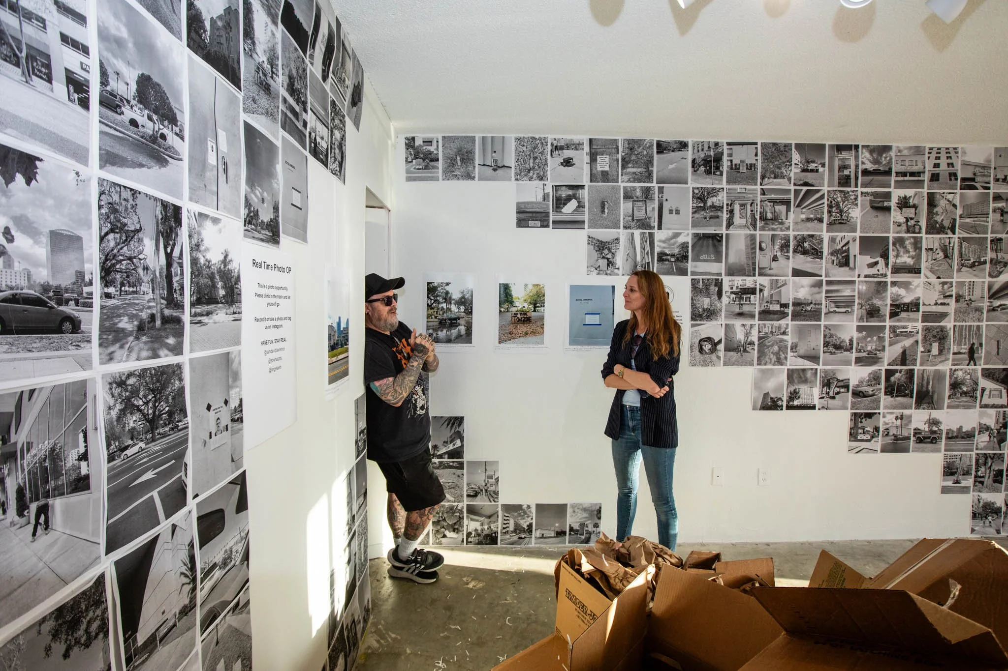 Two people talking in an art gallery with walls covered in black-and-white photos, some color photos, and a sign about real-time photo capture.