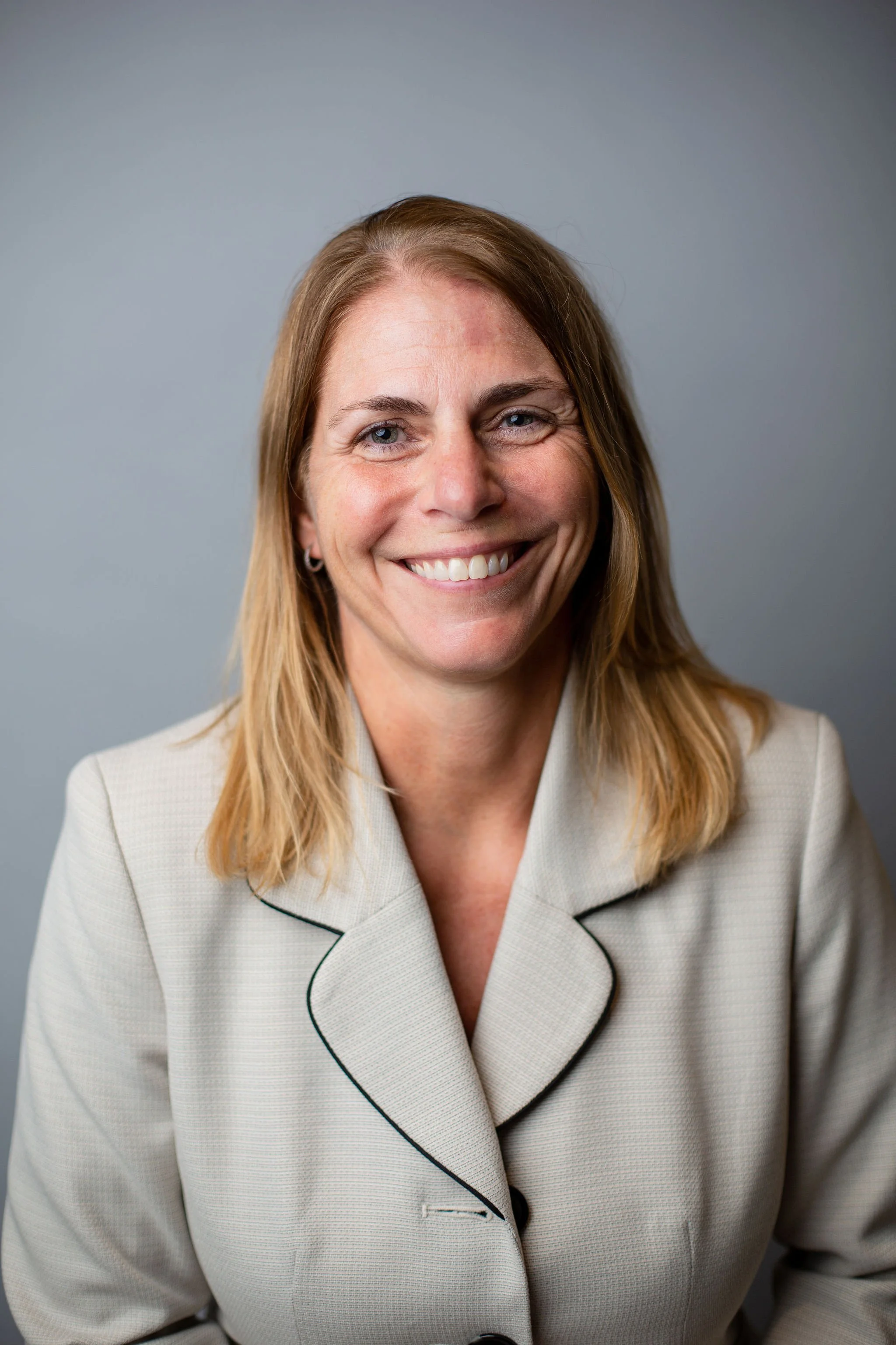 A woman with blonde hair and blue eyes smiling, wearing a beige blazer with black piping, in front of a gray background.