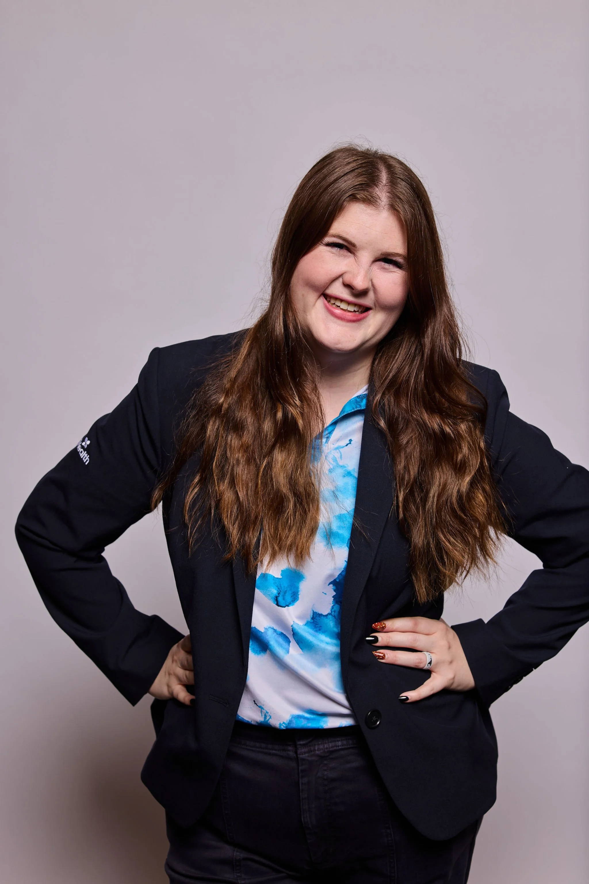 A woman with long brown hair, wearing a dark blazer and a white shirt with blue patterns, posing with her hands on her hips and smiling in front of a plain gray background.