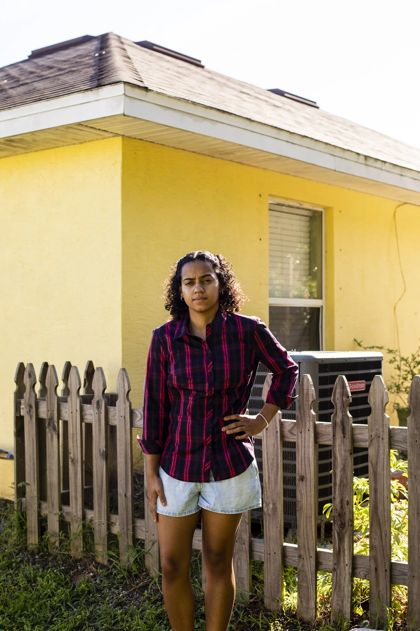 Young woman standing outside in front of a yellow house with a wooden fence, wearing a red and black plaid shirt and shorts.