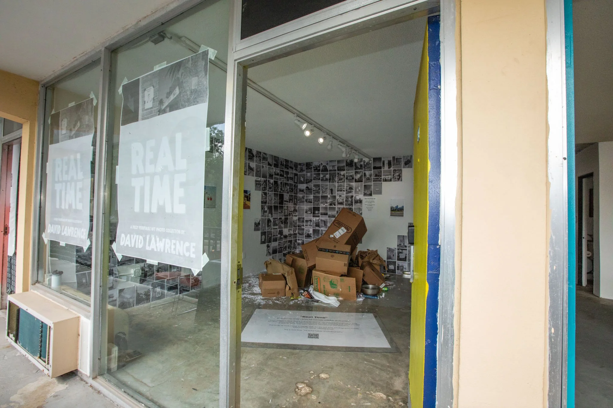 Art gallery exhibit titled "Real Time" by David Lawrence, interior of an art space with cardboard boxes and photo collages on the wall.