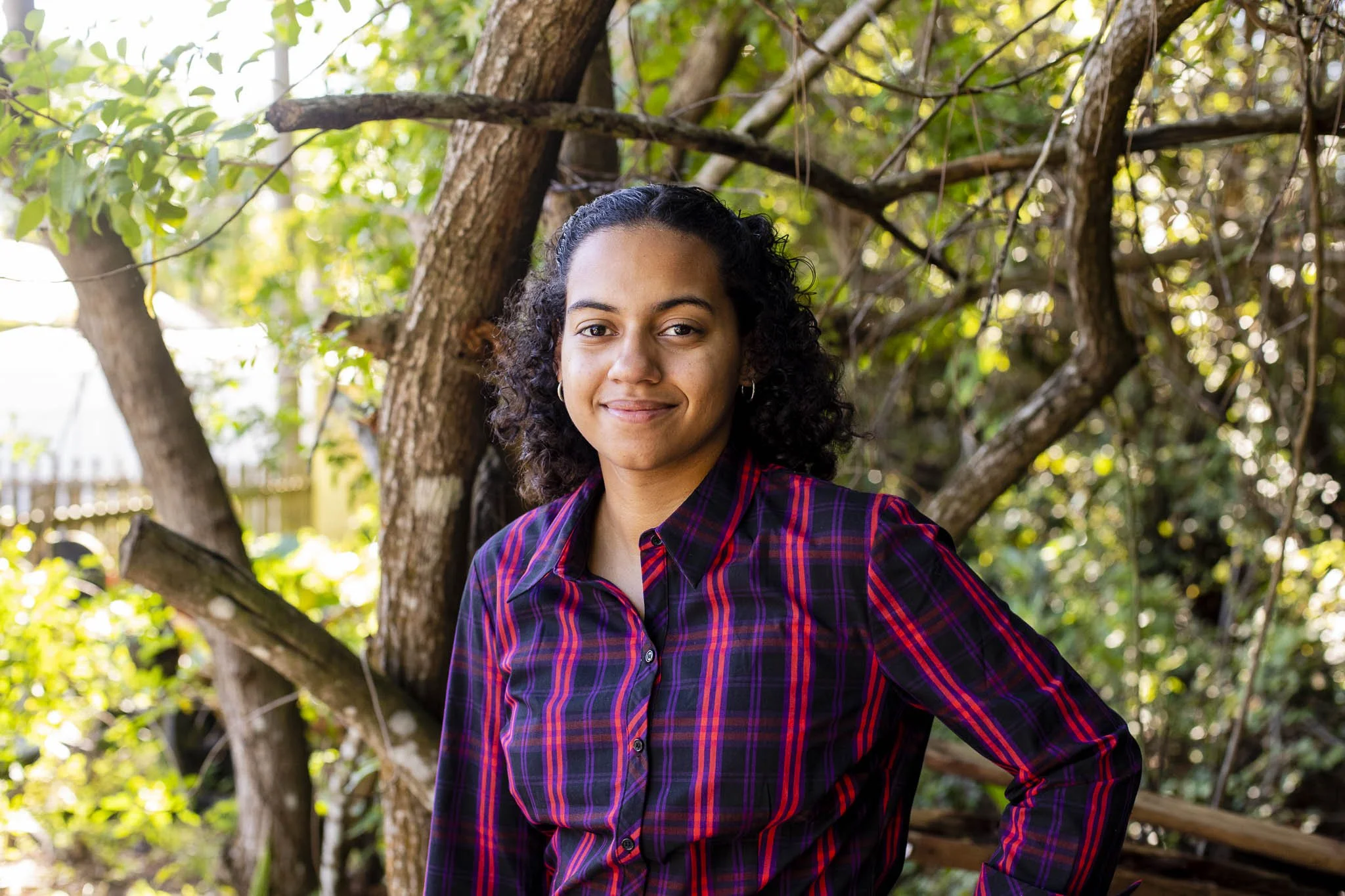 A young woman with dark curly hair, wearing a plaid shirt, standing outdoors in front of a tree with green leaves and branches.
