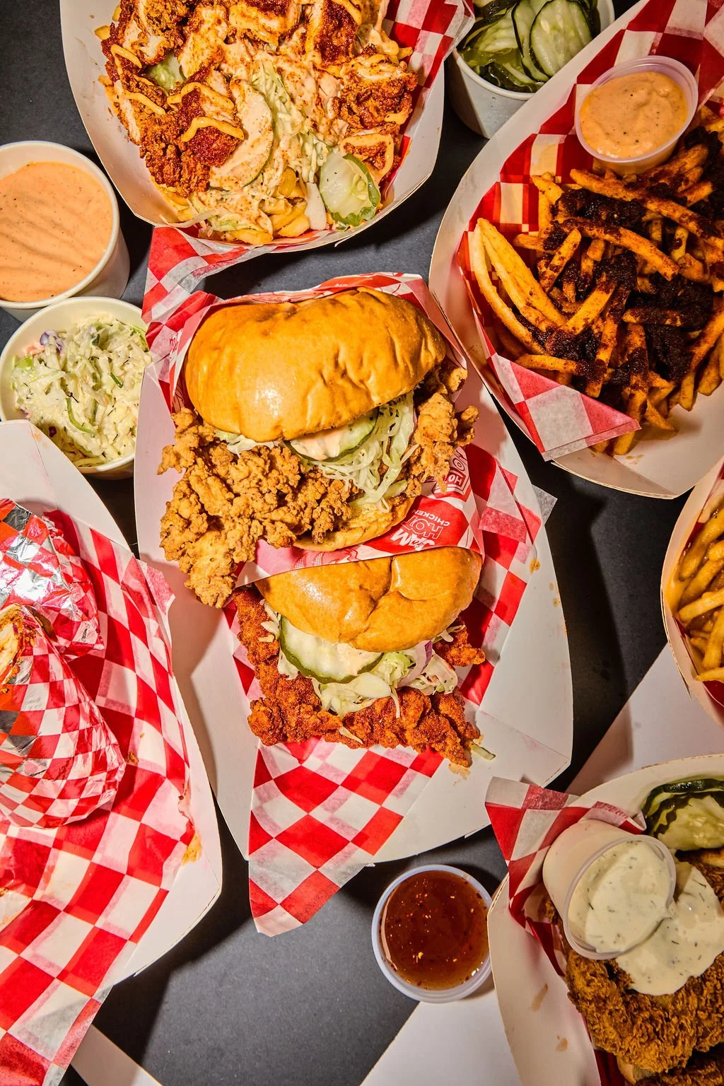 A variety of fast food items including fried chicken sandwiches, French fries, side salads, pickles, and dipping sauces on checkered paper-lined trays.