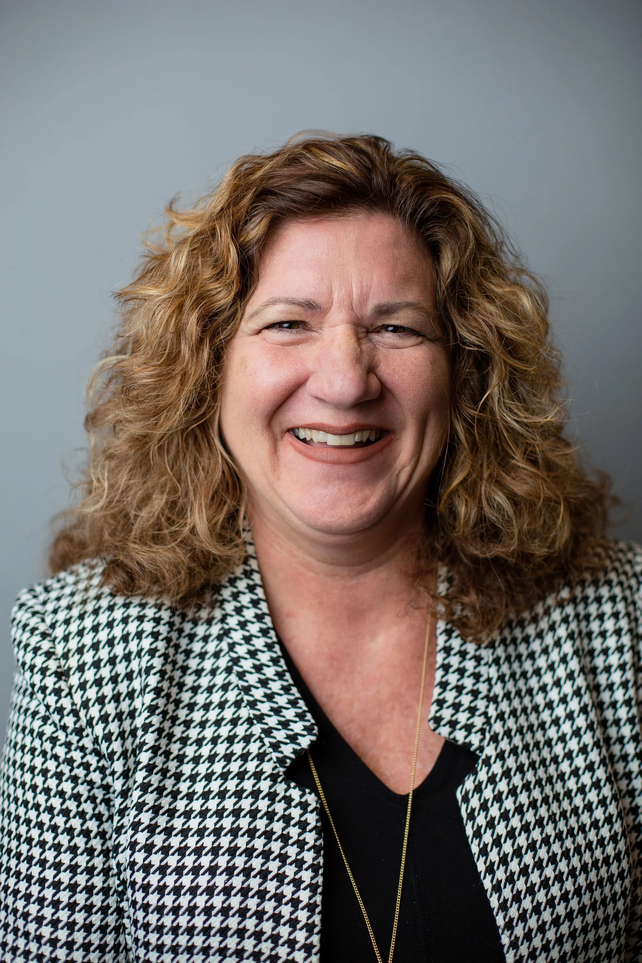 Close-up of middle-aged woman with curly blonde hair, smiling, wearing a black top, a gold necklace, and a houndstooth-patterned blazer against a plain gray background.