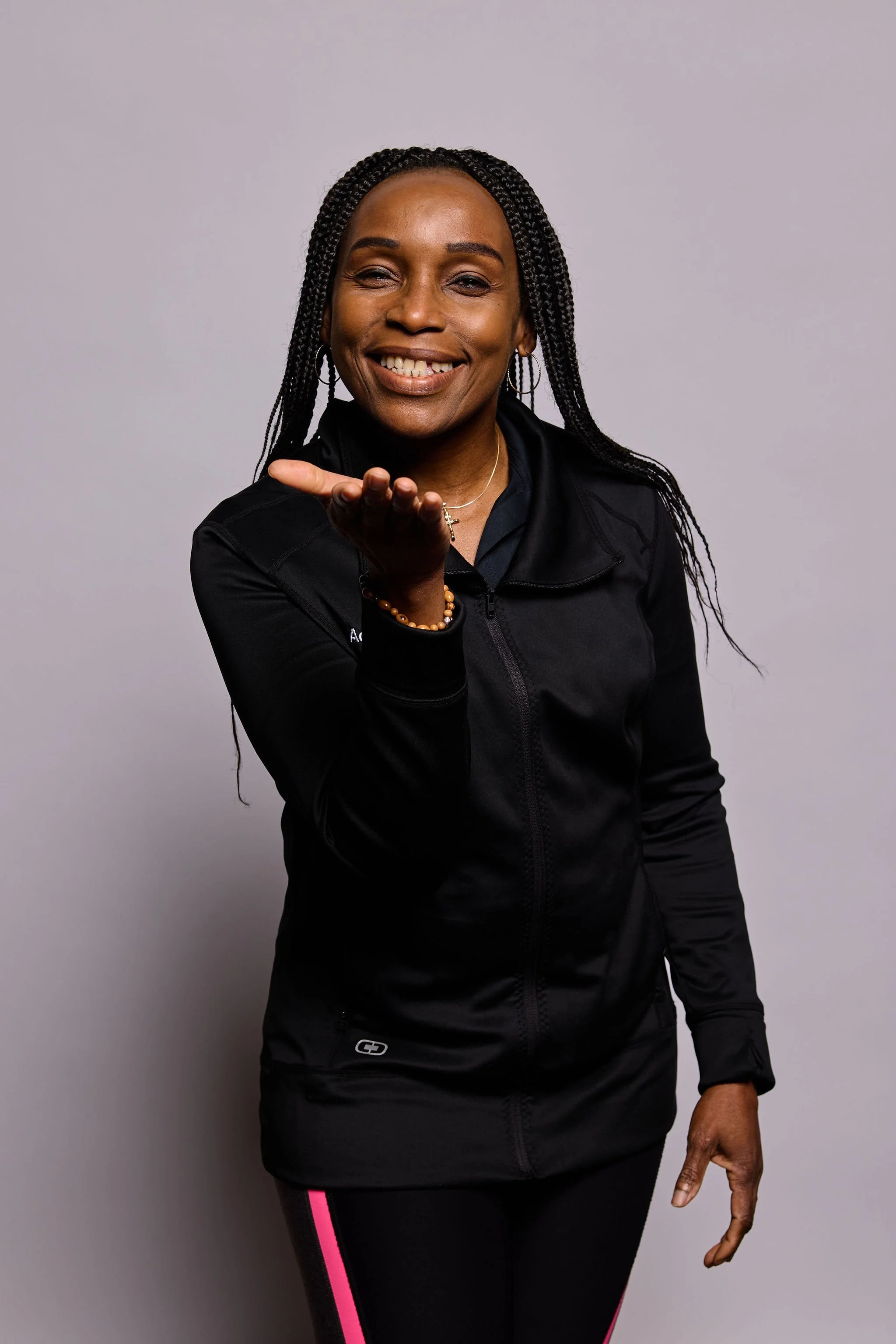 A woman with dark braided hair, wearing a black zip-up jacket and black leggings with pink stripes, smiling and blowing a kiss towards the camera against a plain gray background.